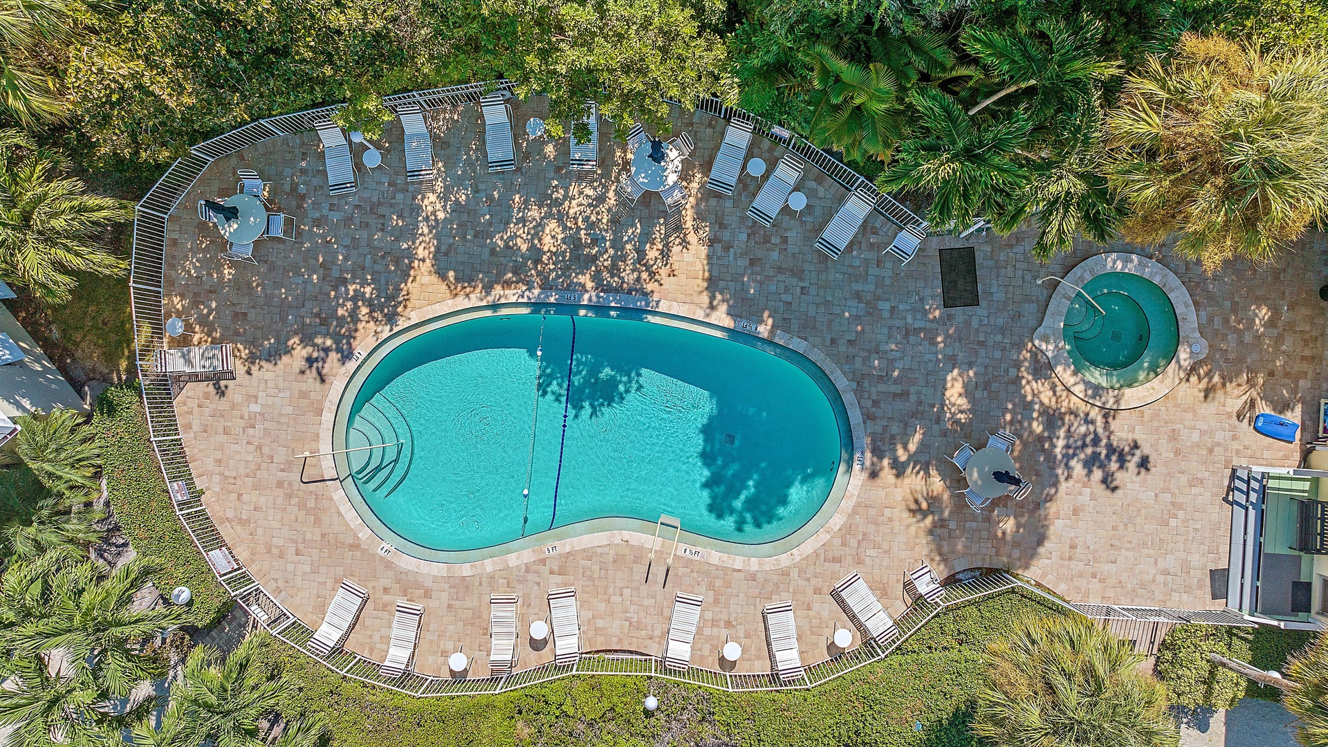 Aerial view of a fenced-in outdoor swimming pool with surrounding lounge chairs, tables, and adjacent hot tub, set amidst greenery.
