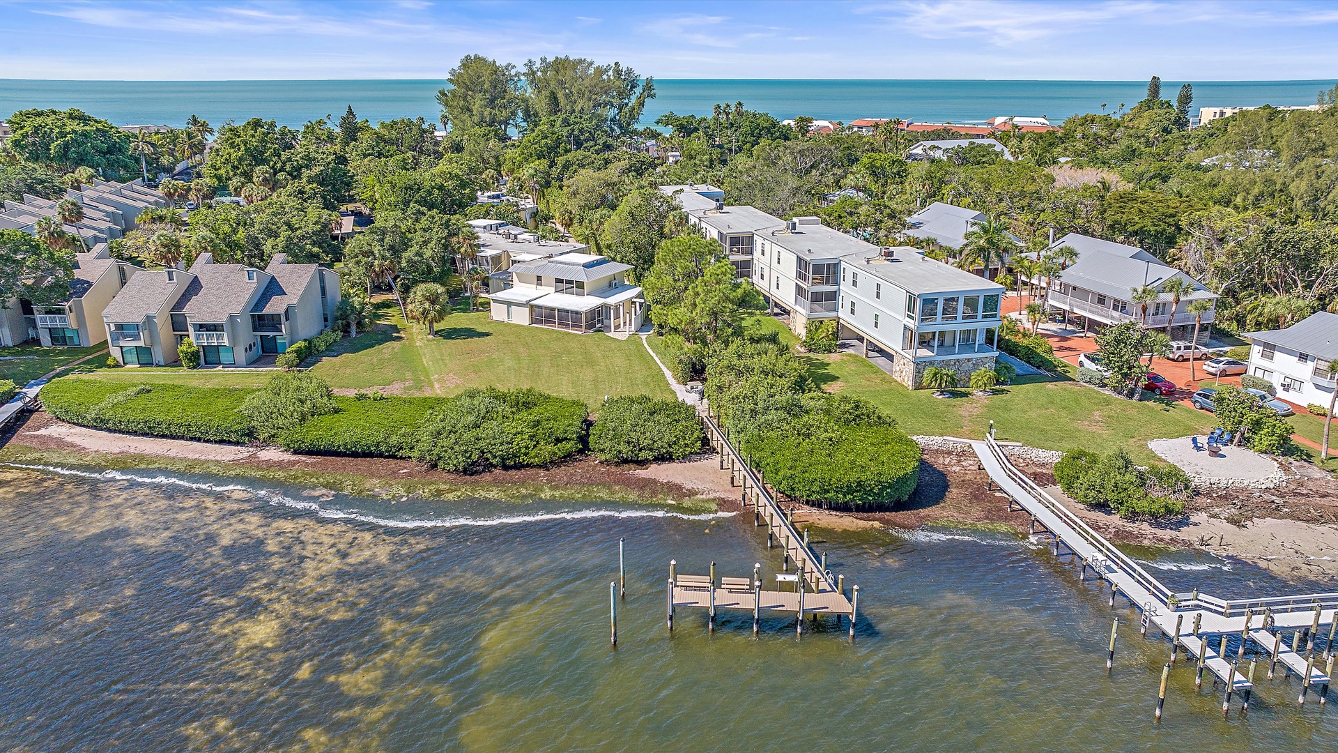 Aerial view of a coastal residential area featuring houses with backyards, a dock, green vegetation, and a shoreline leading to the ocean under a clear blue sky.