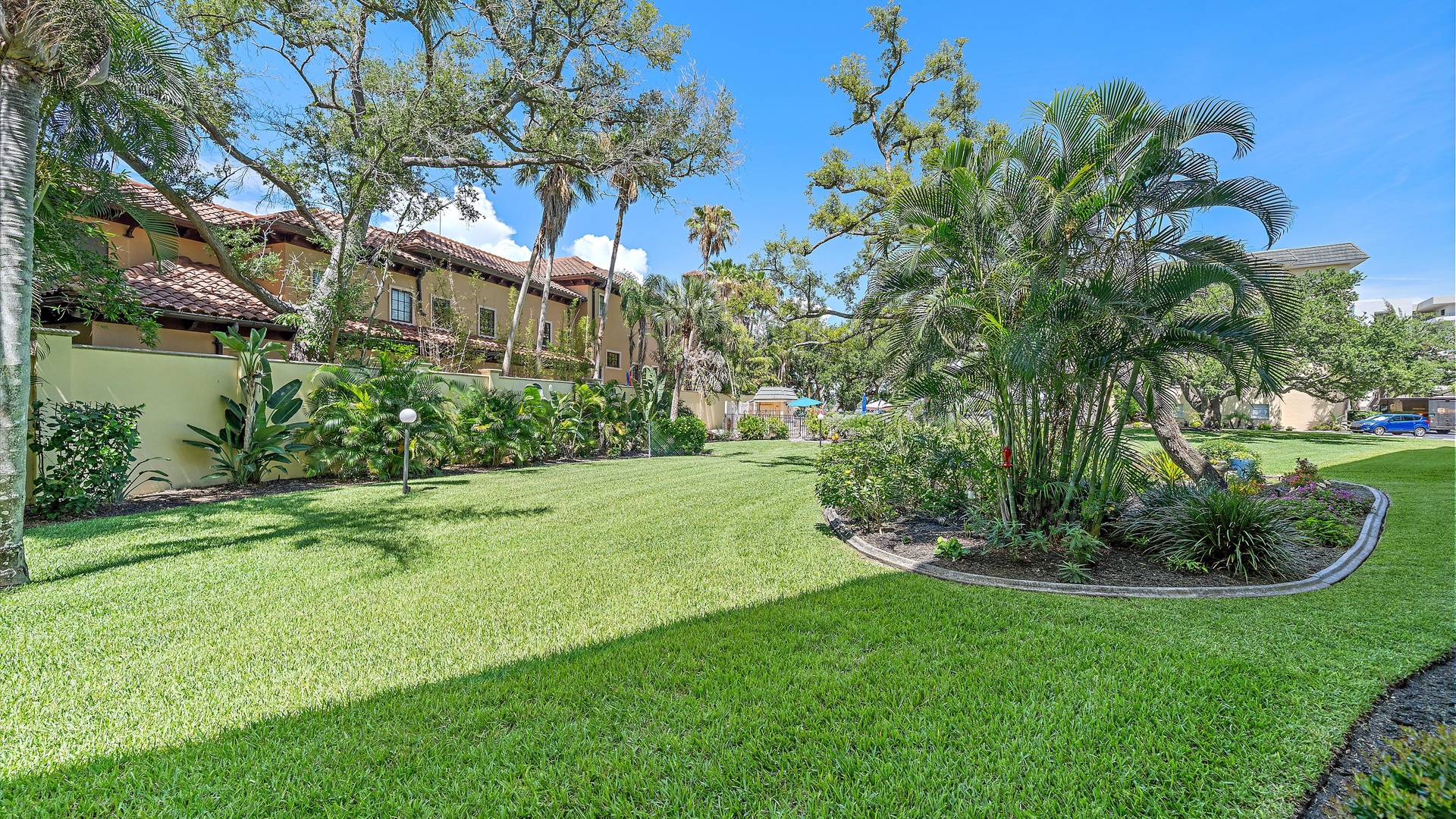 This manicured tropical garden features lush palm trees, mature landscaping, and pristine lawn areas framing the property's Mediterranean-style architecture under sunny skies.