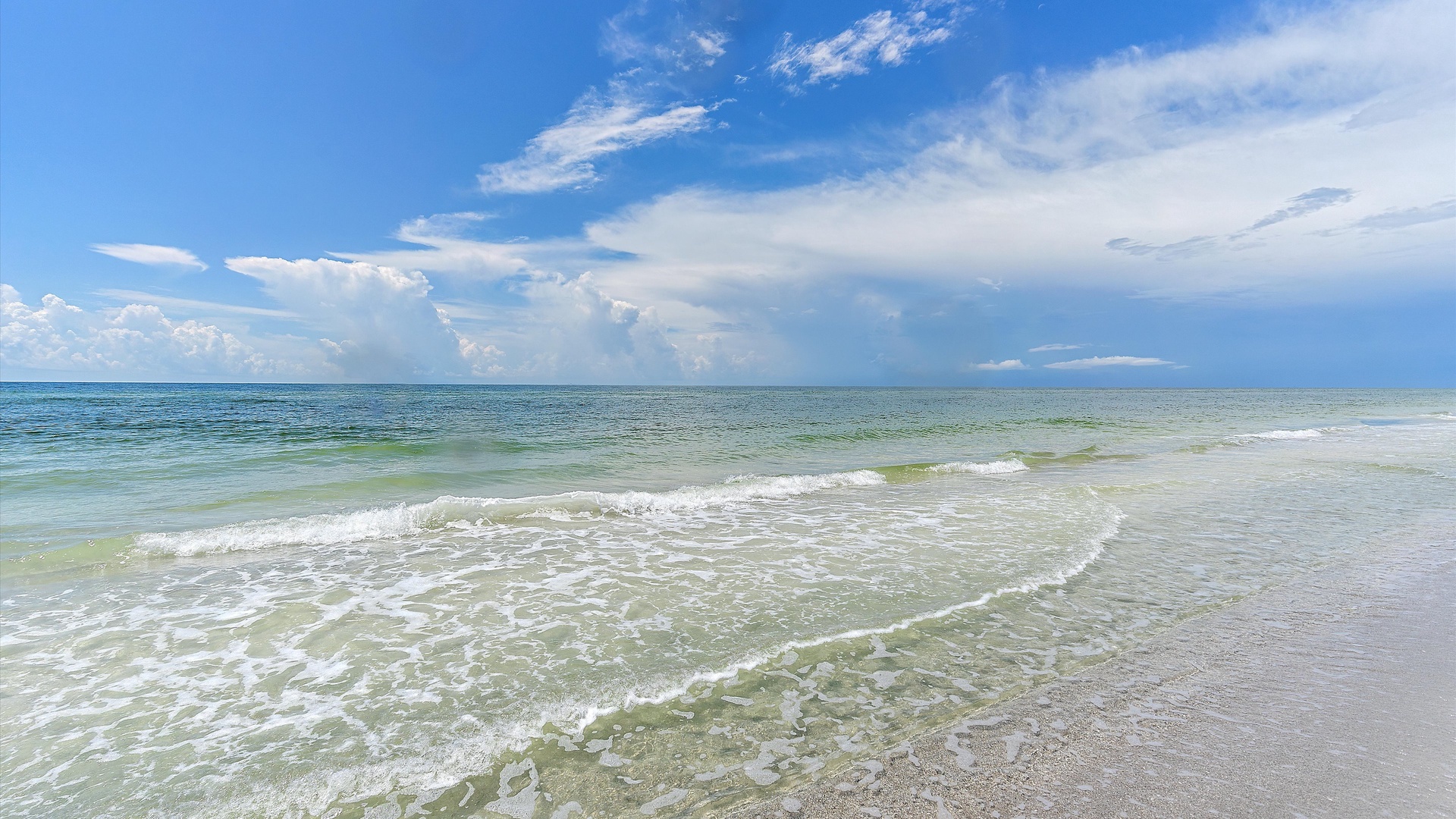 A clear, calm ocean meets a sandy shore under a blue sky with scattered clouds. Gentle waves break at the shoreline.