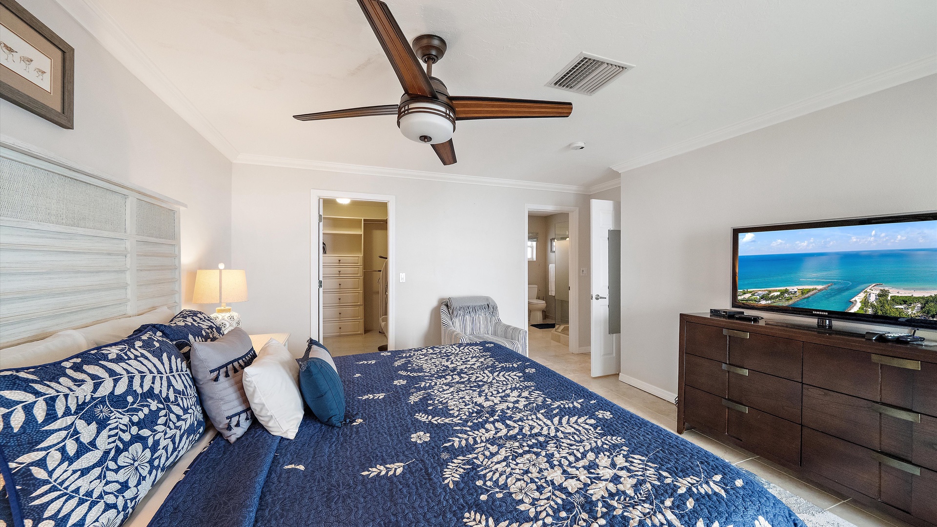 A king-size bed anchors the primary suite, with white furnishings and deep blue accents lending a calm coastal feel. The adjoining bathroom and walk-in closet complete the space.