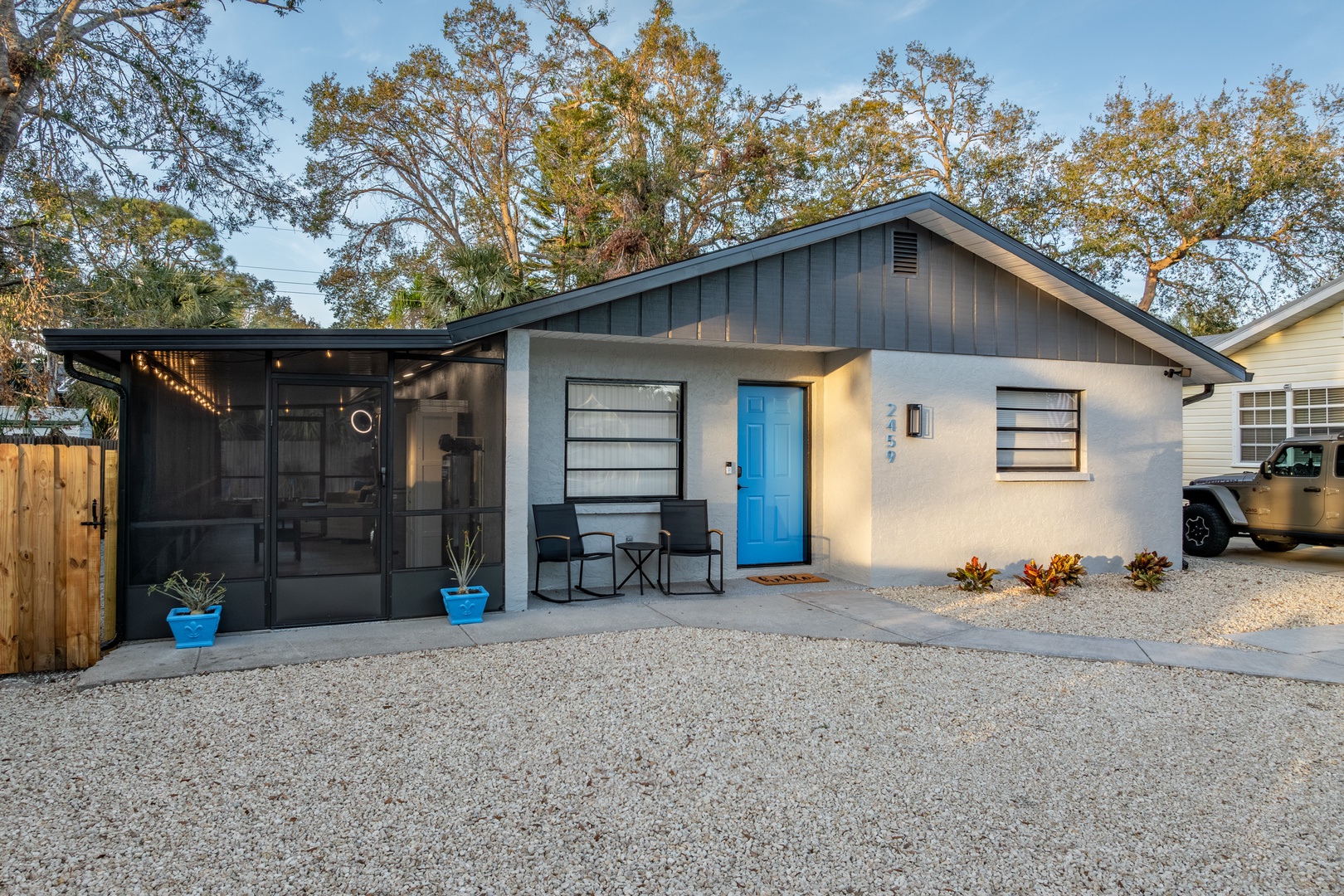 Modern cottage with bright blue door and screened porch, surrounded by mature trees and private gravel parking.