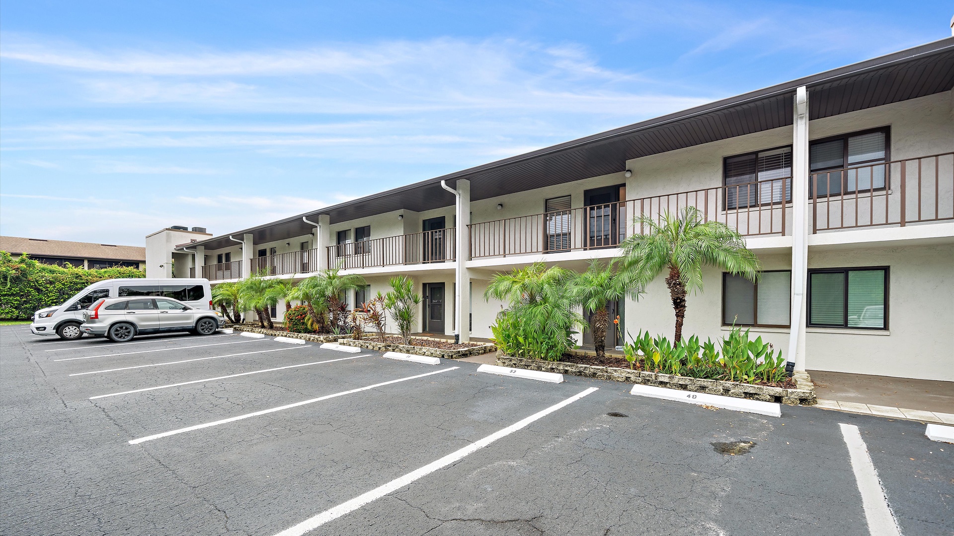 The two-story property building features convenient parking spaces and tropical plantings under a bright blue sky, creating a welcoming arrival experience.