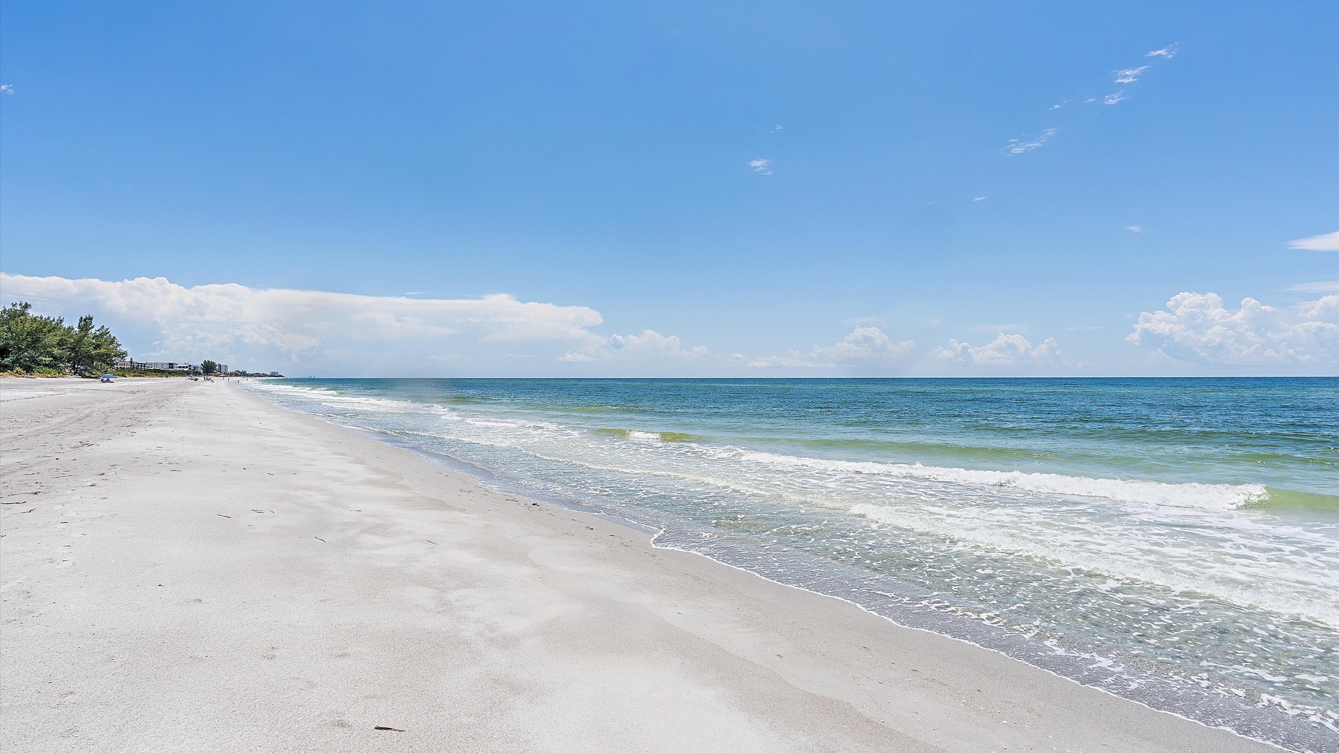 A pristine beach with white sand extends along the shoreline under a clear blue sky. Gentle waves lap at the shore, and a line of green trees is visible in the distance.