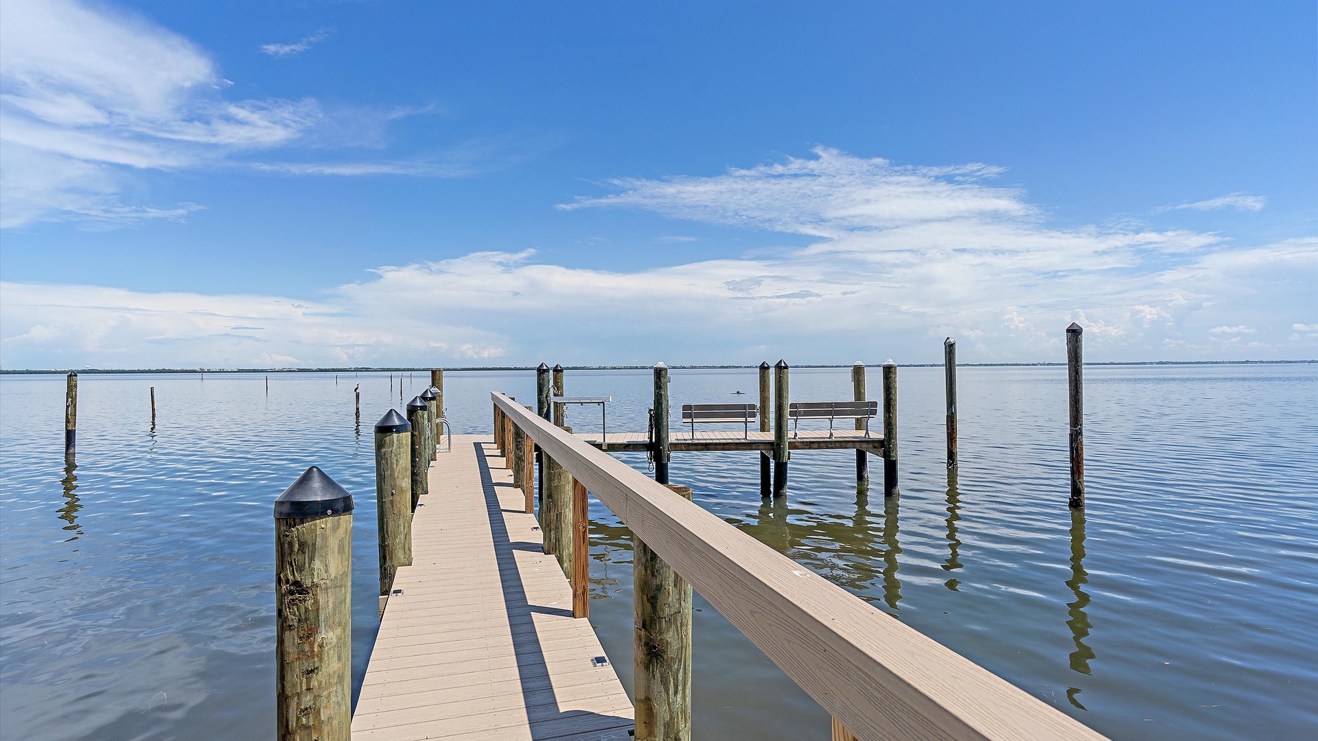 A wooden pier extends over calm water, leading to a small platform with a bench under a clear blue sky. Multiple wooden pilings are visible in the water.