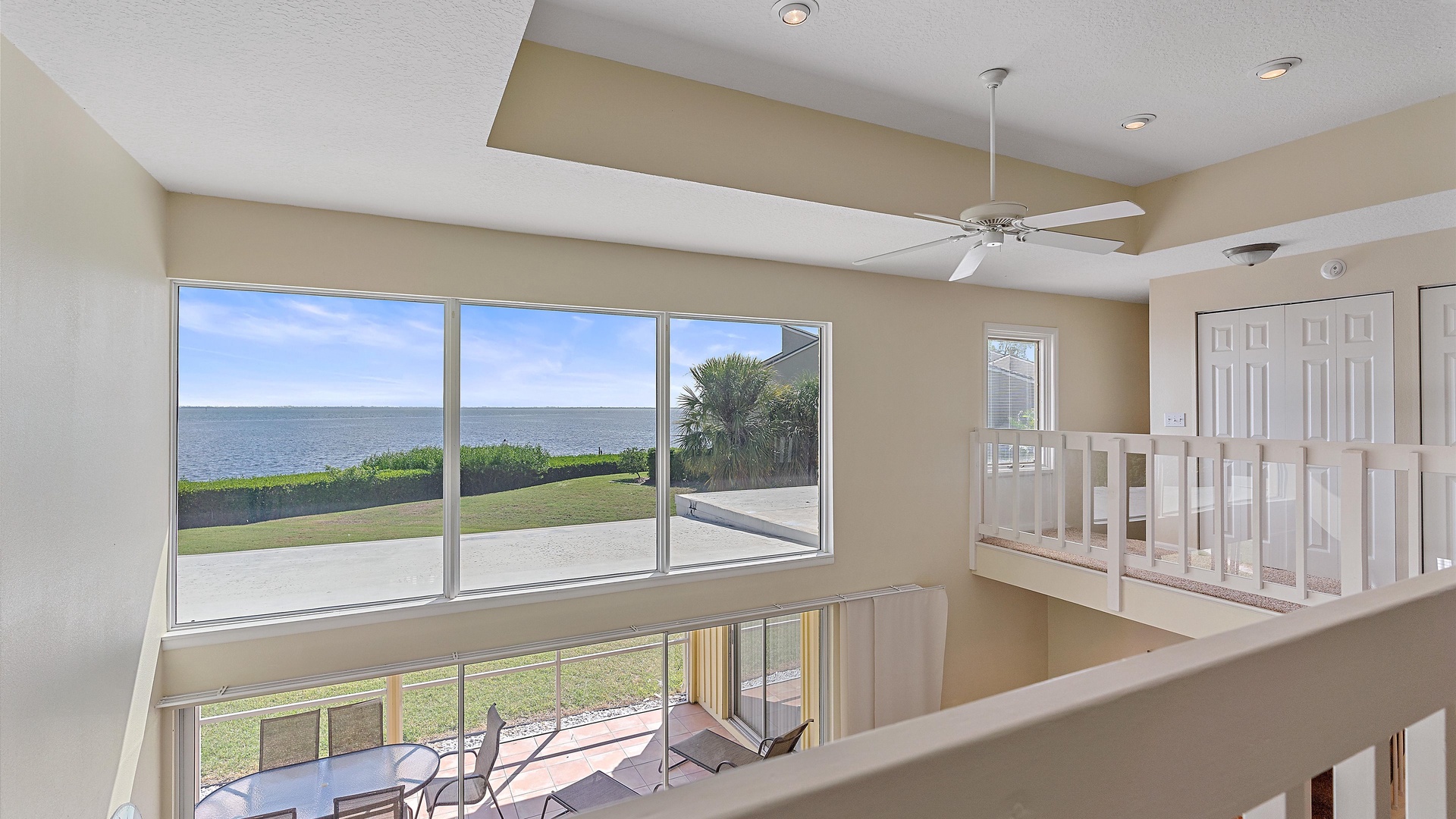 A two-story room with large windows overlooking a grassy area and a body of water. There is a table and chairs on a patio outside. The ceiling has recessed lighting and a ceiling fan.