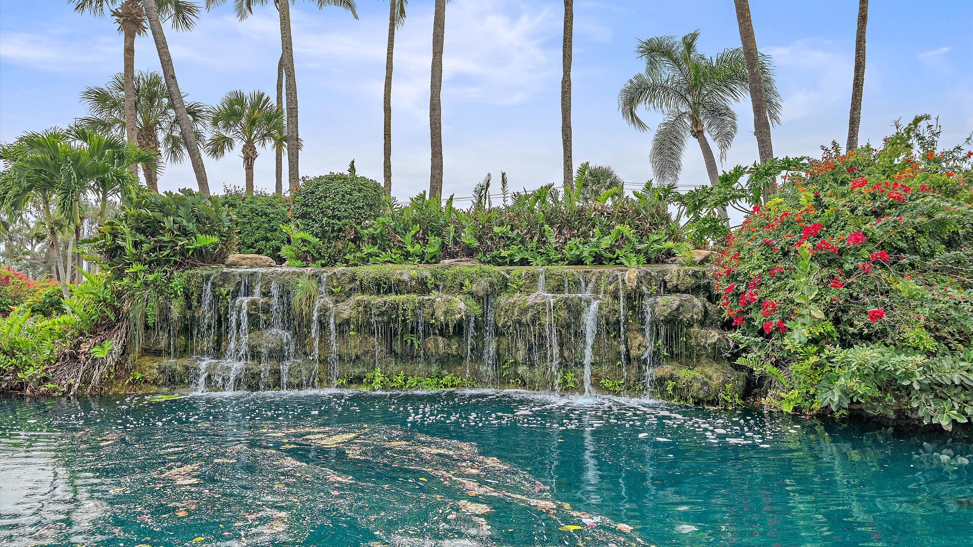 A stunning tropical swimming pool featuring a dramatic rock waterfall surrounded by lush palm trees and vibrant flowering plants in the area.