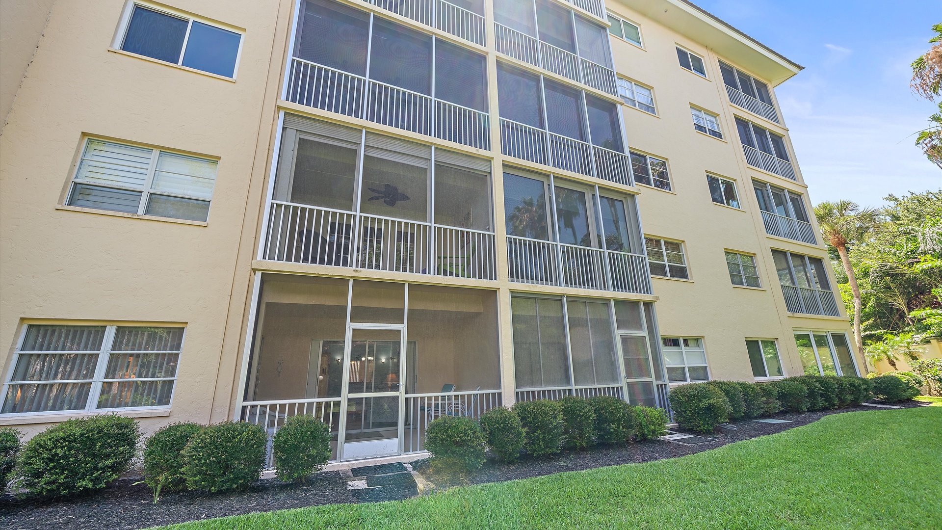 Multi-story residential building featuring screened balconies, manicured landscaping, and surrounded by lush tropical vegetation under clear blue skies.