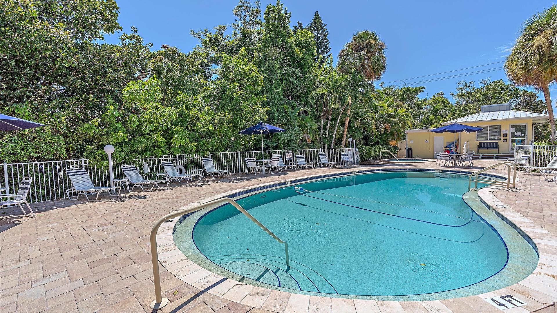Outdoor pool area with lounge chairs and umbrellas set up around the perimeter. Surrounded by dense greenery and trees. Adjacent building with a covered seating area. Clear, sunny day.