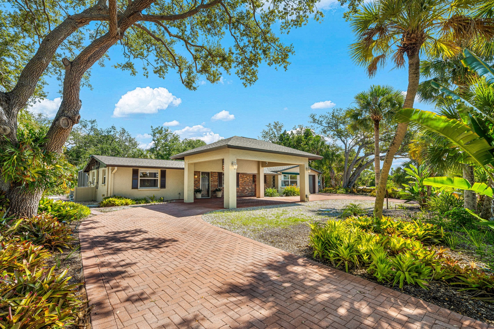 A charming property set amid lush tropical landscaping, featuring a covered carport and easy access along a paved brick walkway.