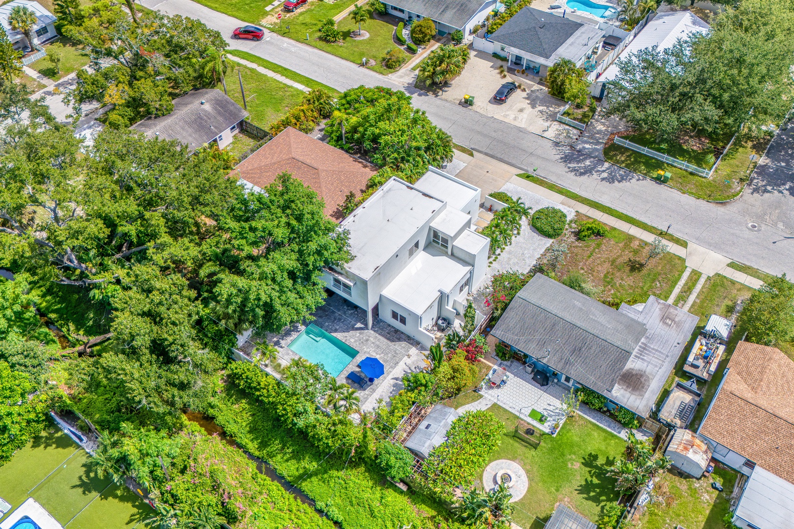 Aerial view of a modern white property nestled in a lush residential neighborhood, featuring a private pool and abundant greenery.