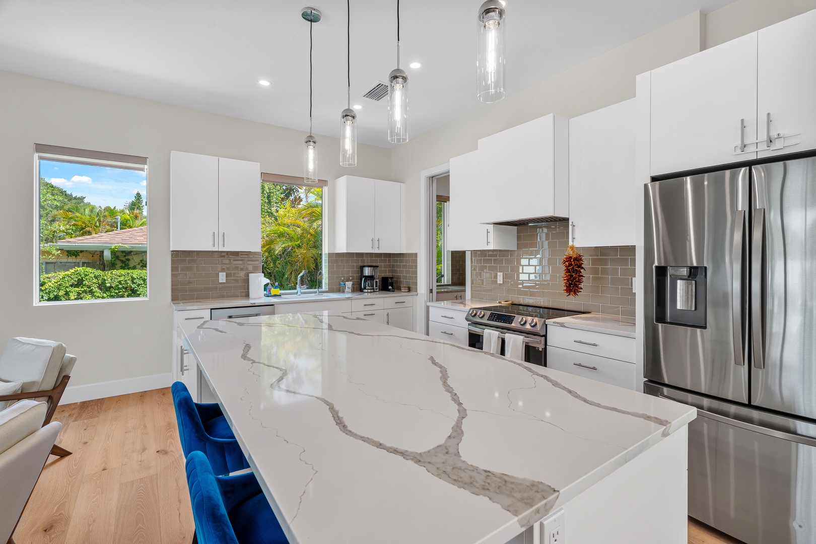 Prepare your favorite meals in this pristine kitchen featuring gleaming marble counters, stainless appliances, and bright natural light streaming through generous windows overlooking lush greenery.