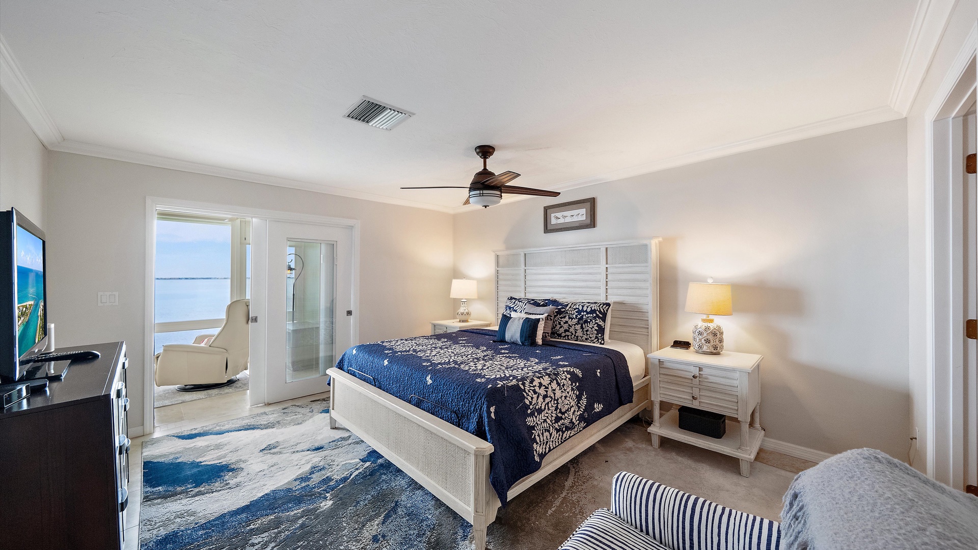 A restful king bedroom in navy, white, and soft coastal neutrals. Doors open to the enclosed sunroom, connecting this comfortable retreat with the penthouse’s signature waterfront setting.