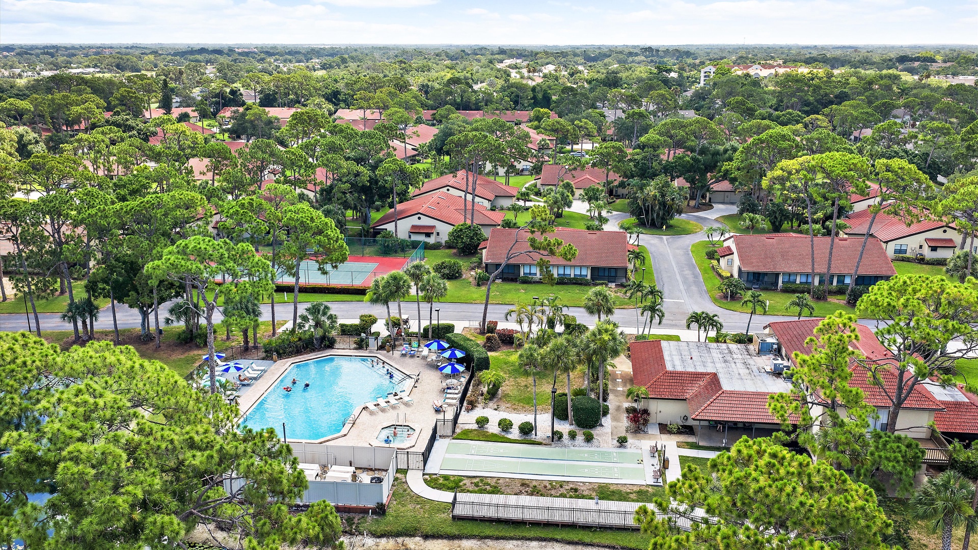 Aerial view of a tree-lined residential community featuring a sparkling pool and red-tiled roof homes surrounded by lush Florida landscaping and winding neighborhood streets.