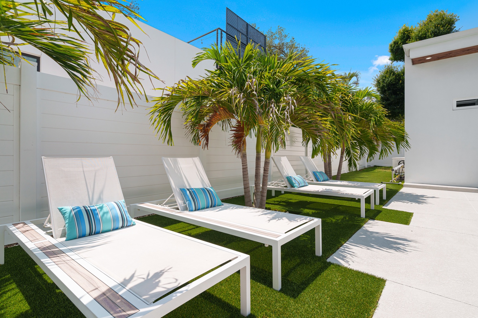 Modern lounge area featuring sleek white sun loungers, vibrant palm trees, and lush green lawn under clear blue skies—an ideal spot to unwind and soak up the sun.