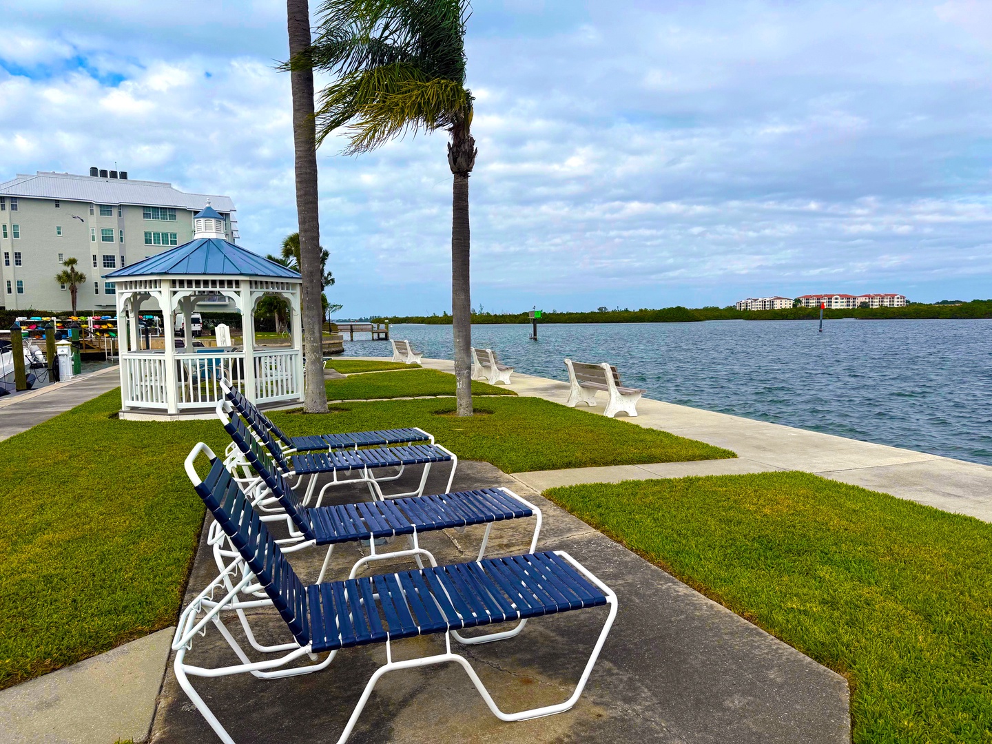 Waterfront lounge chairs and gazebo overlooking the bay, with palm trees and property buildings visible in the distance.