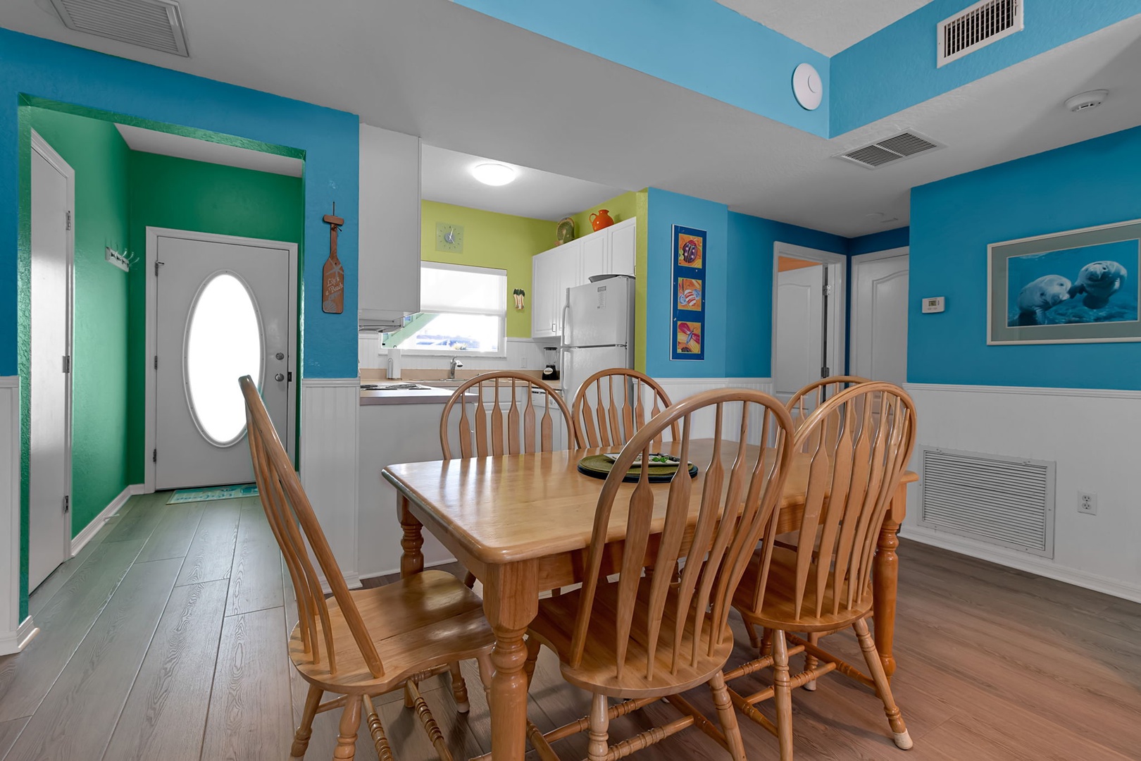 Bright, open-concept living with cheerful teal walls and natural light streaming in—perfect for gathering around the wooden dining table with your crew after a day at the beach.