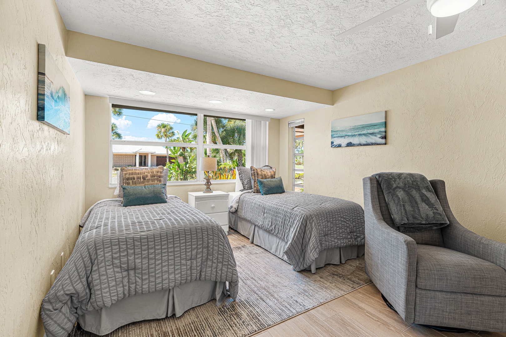 Your coastal retreat awaits with plush twin beds dressed in soothing gray tones, plus a cozy armchair to sink into. Bright windows frame tropical palms outside, filling the room with natural light and easy beach vibes.