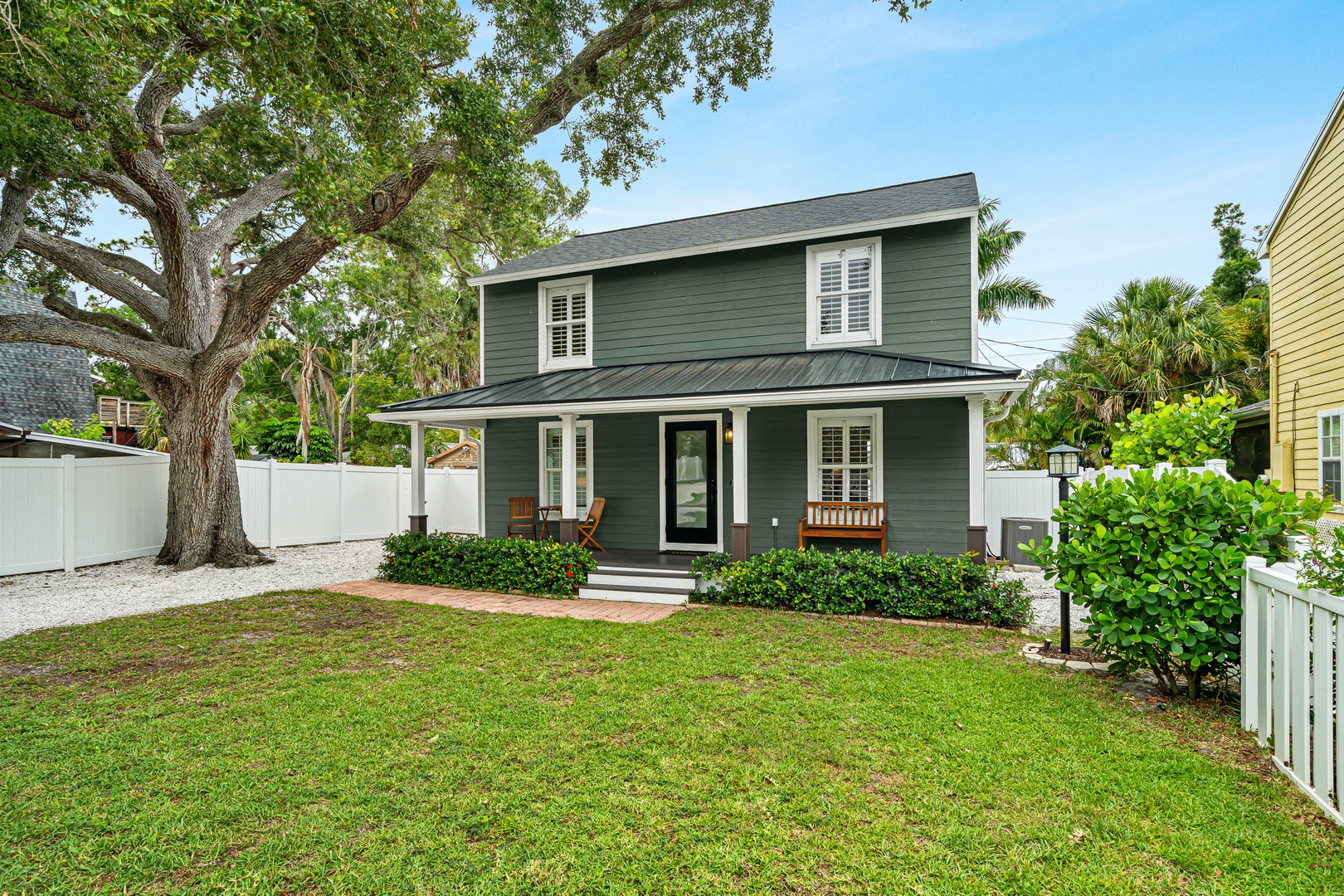 A charming two-story coastal retreat with a welcoming front porch, surrounded by lush tropical greenery and mature shade trees in a peaceful residential neighborhood.