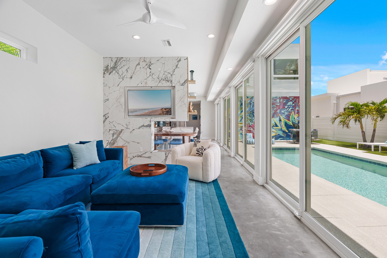 Sink into plush seating as bright sunshine streams through floor-to-ceiling glass, framing your private pool and tropical palms—a seamless blend of luxury indoors and paradise just steps away.
