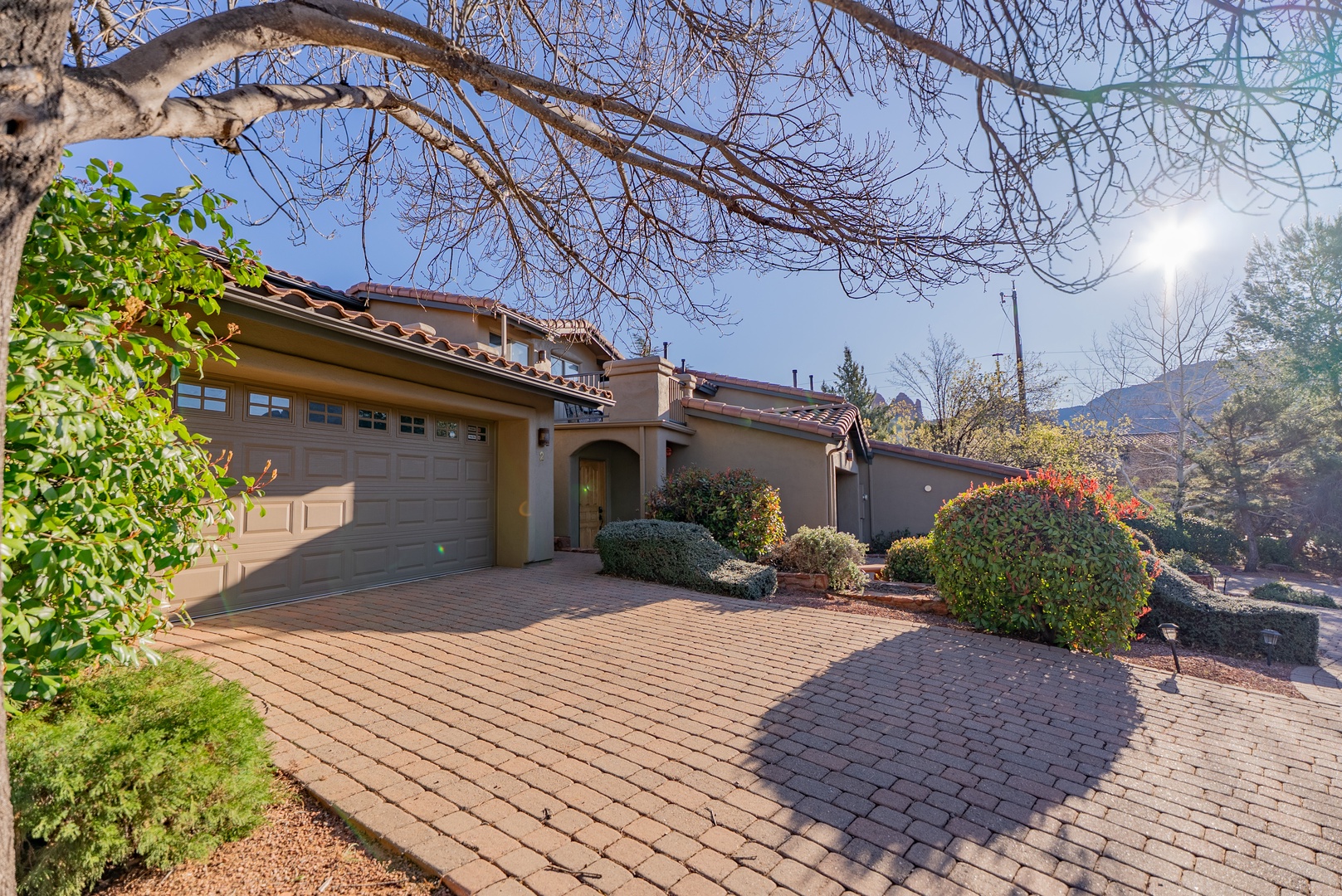 Elegant Mediterranean-style home with tile roof and brick driveway, surrounded by mature landscaping and mountain views.