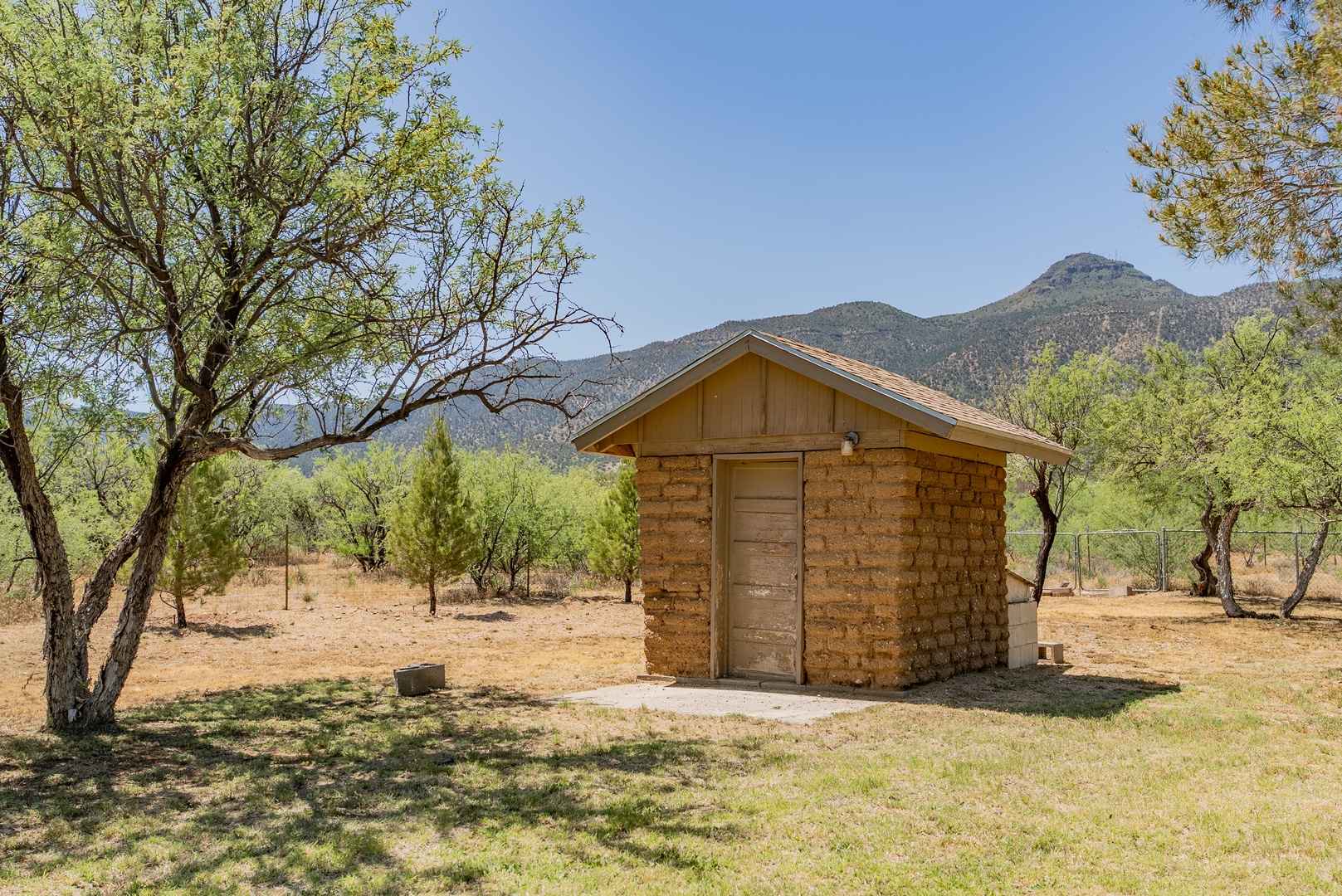 A rustic stone cabin nestled beneath towering mountains, surrounded by desert trees and endless blue skies in this peaceful natural setting.