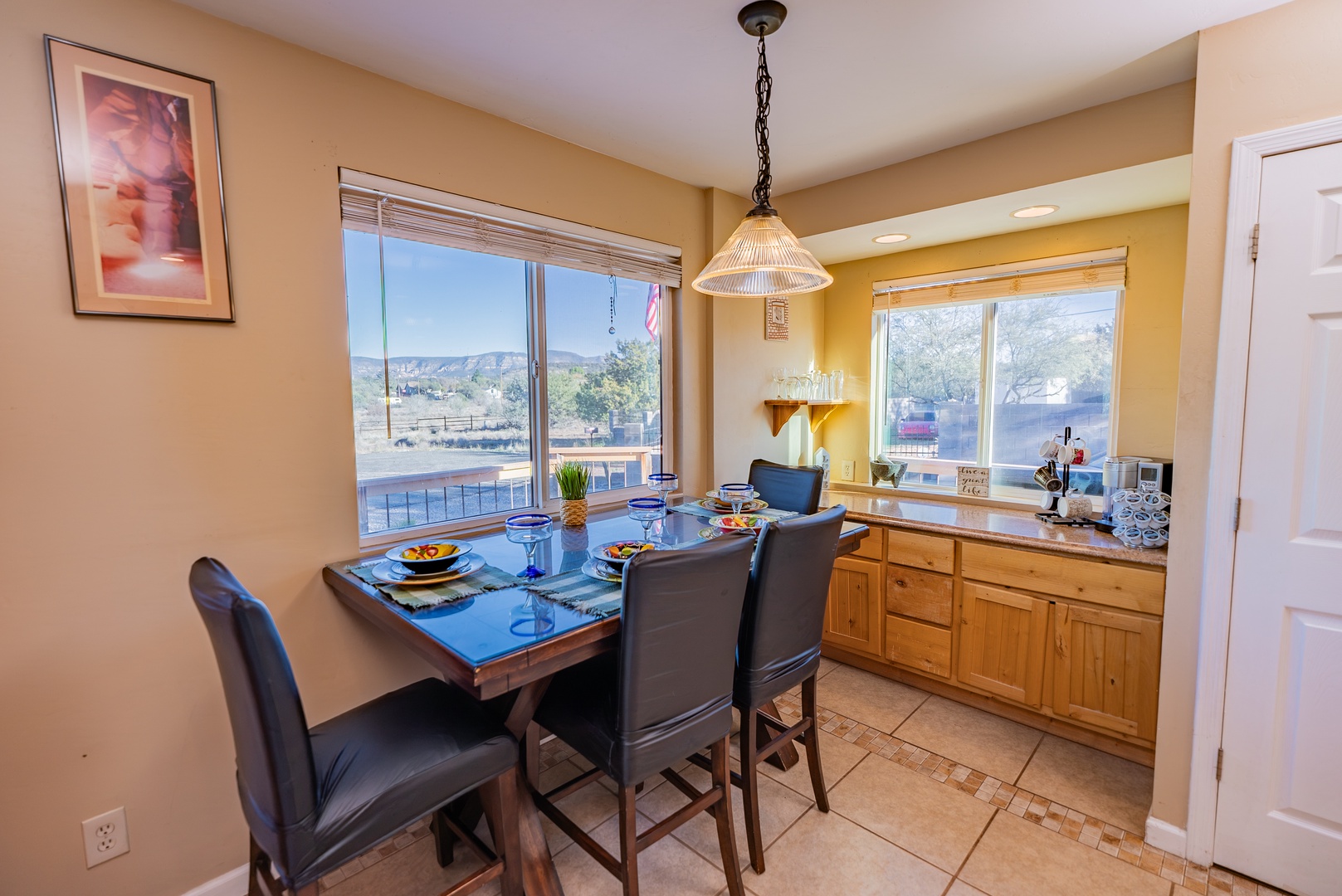 Your elegant dining space features a beautifully set table for four, warm wood cabinetry, and abundant natural light streaming through large windows for memorable meals together.