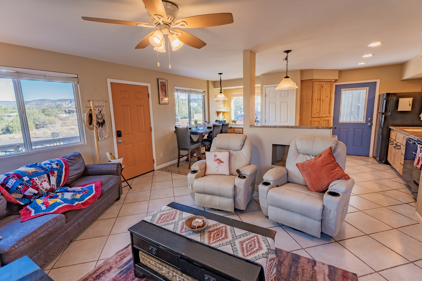 Relax in your spacious open-concept living area featuring comfortable seating, ceiling fan, and panoramic windows showcasing the scenic landscape beyond.