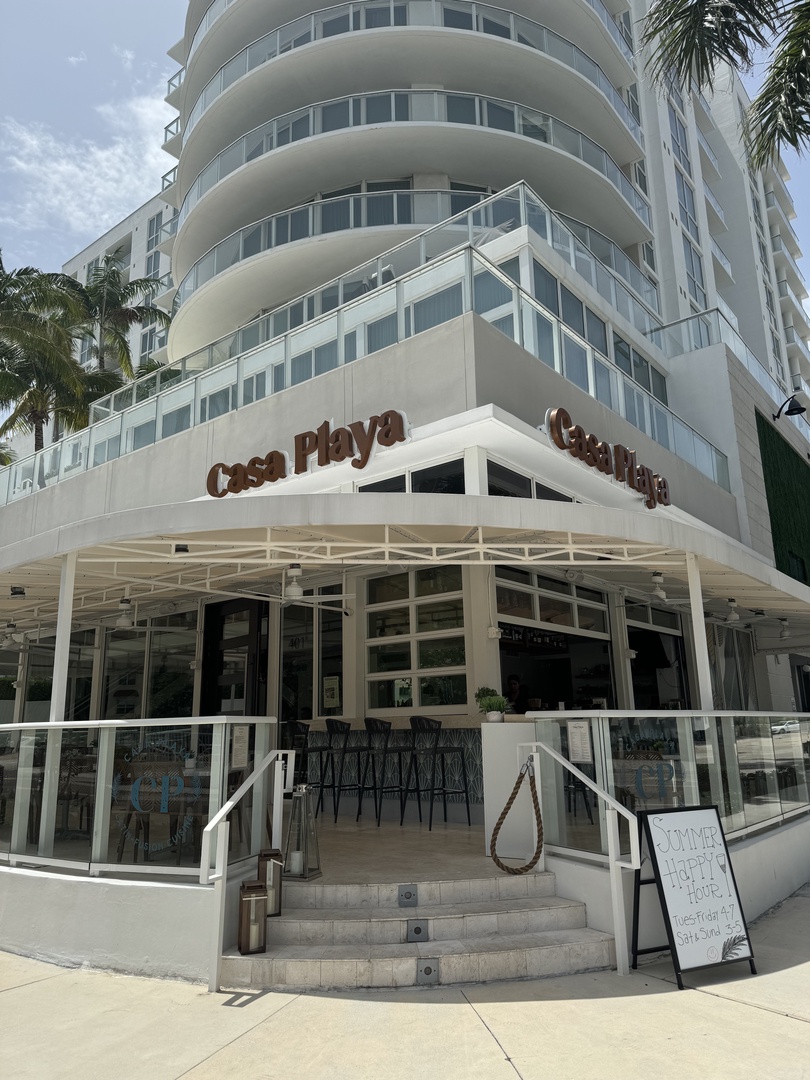 Modern white building with distinctive curved balconies and signage marking the property entrance, situated in a tropical urban setting with palm trees.