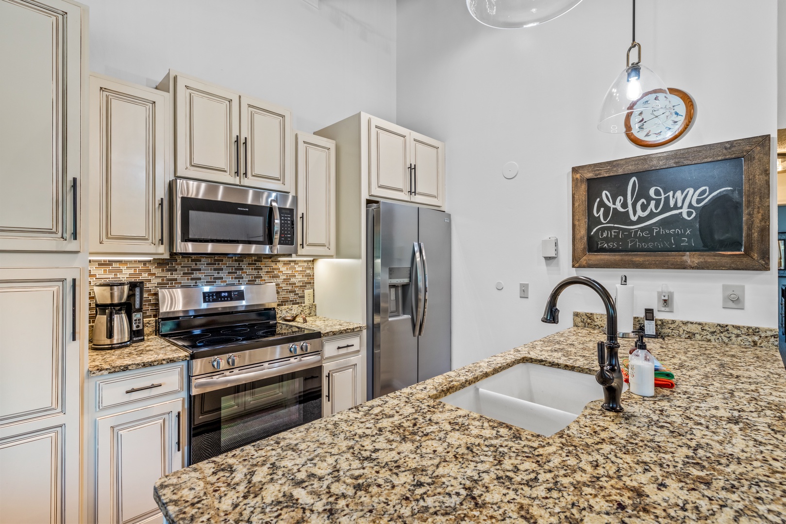 Your fully equipped kitchen invites you to prepare meals with ease, featuring modern stainless steel appliances, granite countertops, and a welcoming chalkboard message—all set under bright pendant lighting.