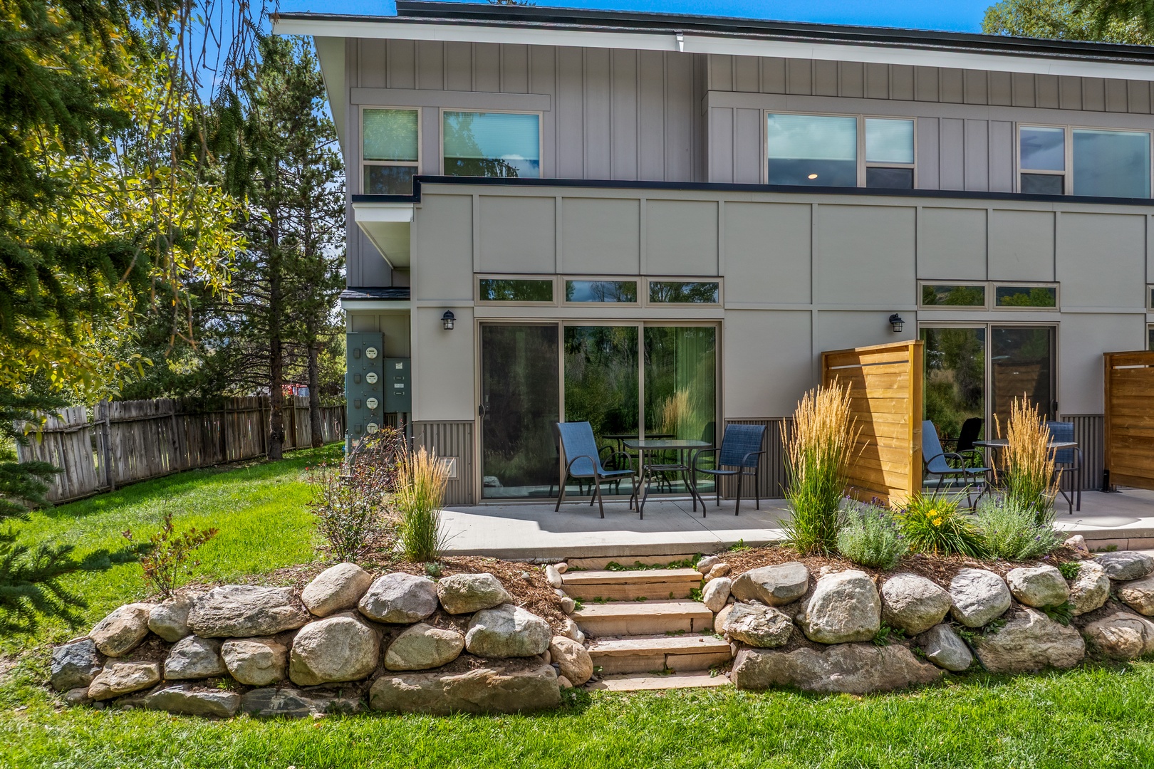 Modern property exterior framed by mature trees and lush green lawn, with stone-bordered garden beds leading to private patio spaces.