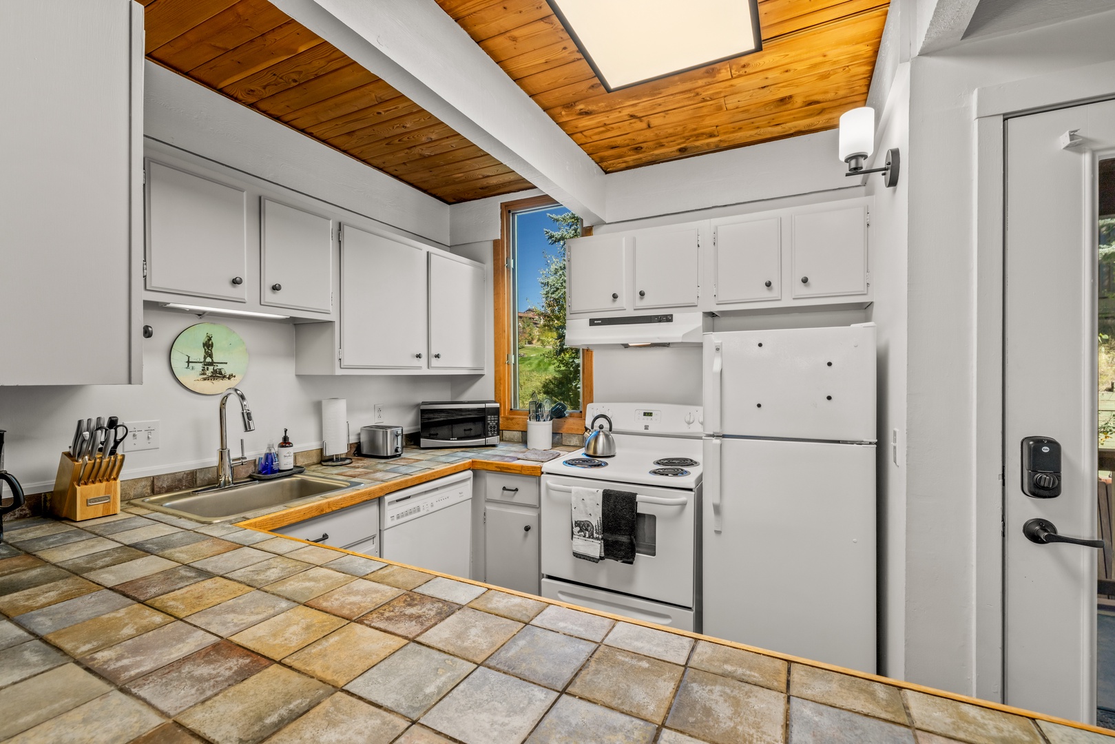 Your fully-equipped kitchen features warm wood-beamed ceilings, crisp white cabinetry, and abundant natural light streaming through the window, creating a welcoming space to cook and gather.