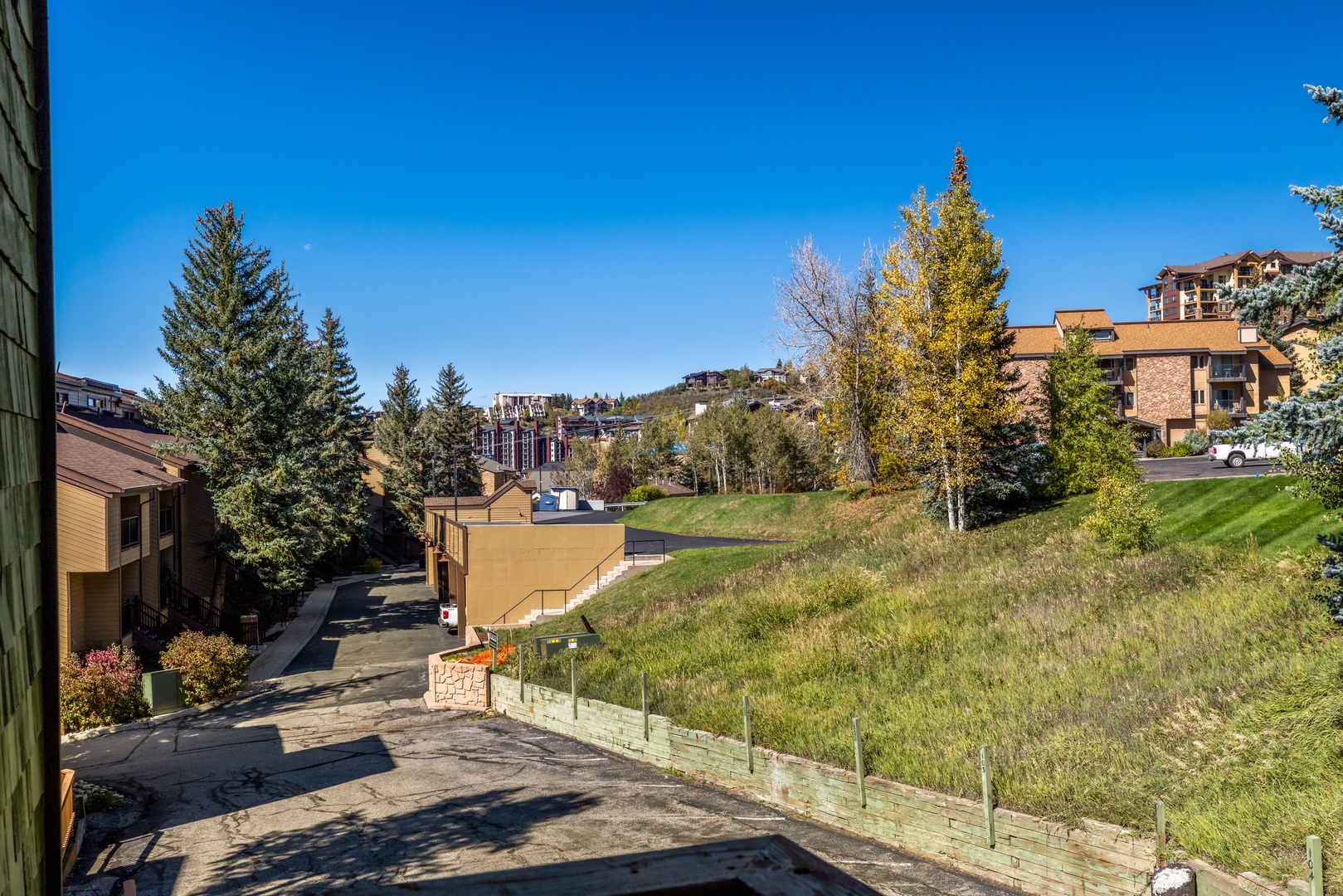 The property sits in a quiet mountain neighborhood surrounded by evergreens and aspens, with nearby lodges and buildings visible on the hillside. A peaceful street leads to the entrance, offering convenient access to the surrounding area.
