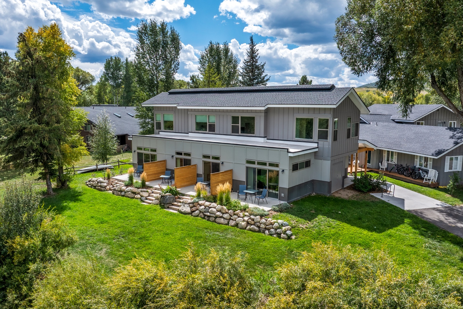 A modern vacation rental sits nestled among mature trees, featuring a spacious lawn and thoughtfully landscaped stone pathways leading to a private outdoor patio—your peaceful mountain retreat awaits.