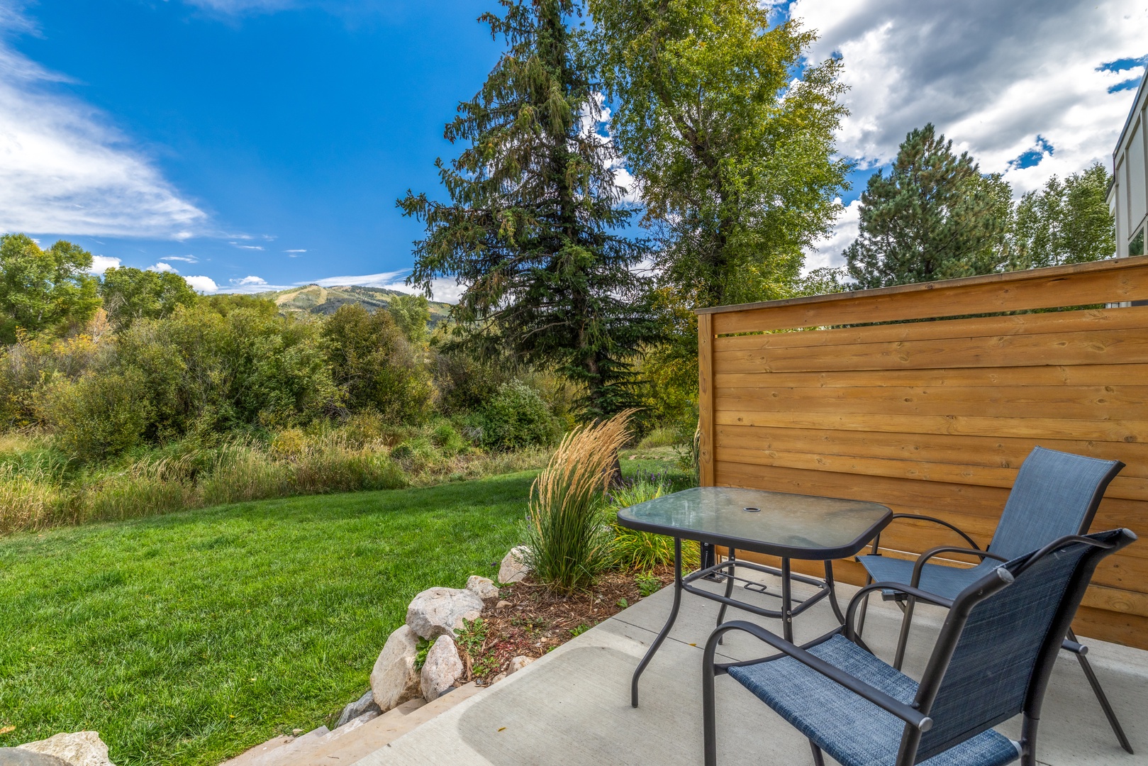 Your private patio opens to a serene mountain backdrop, where lush trees frame distant peaks and a manicured lawn invites quiet moments outdoors.