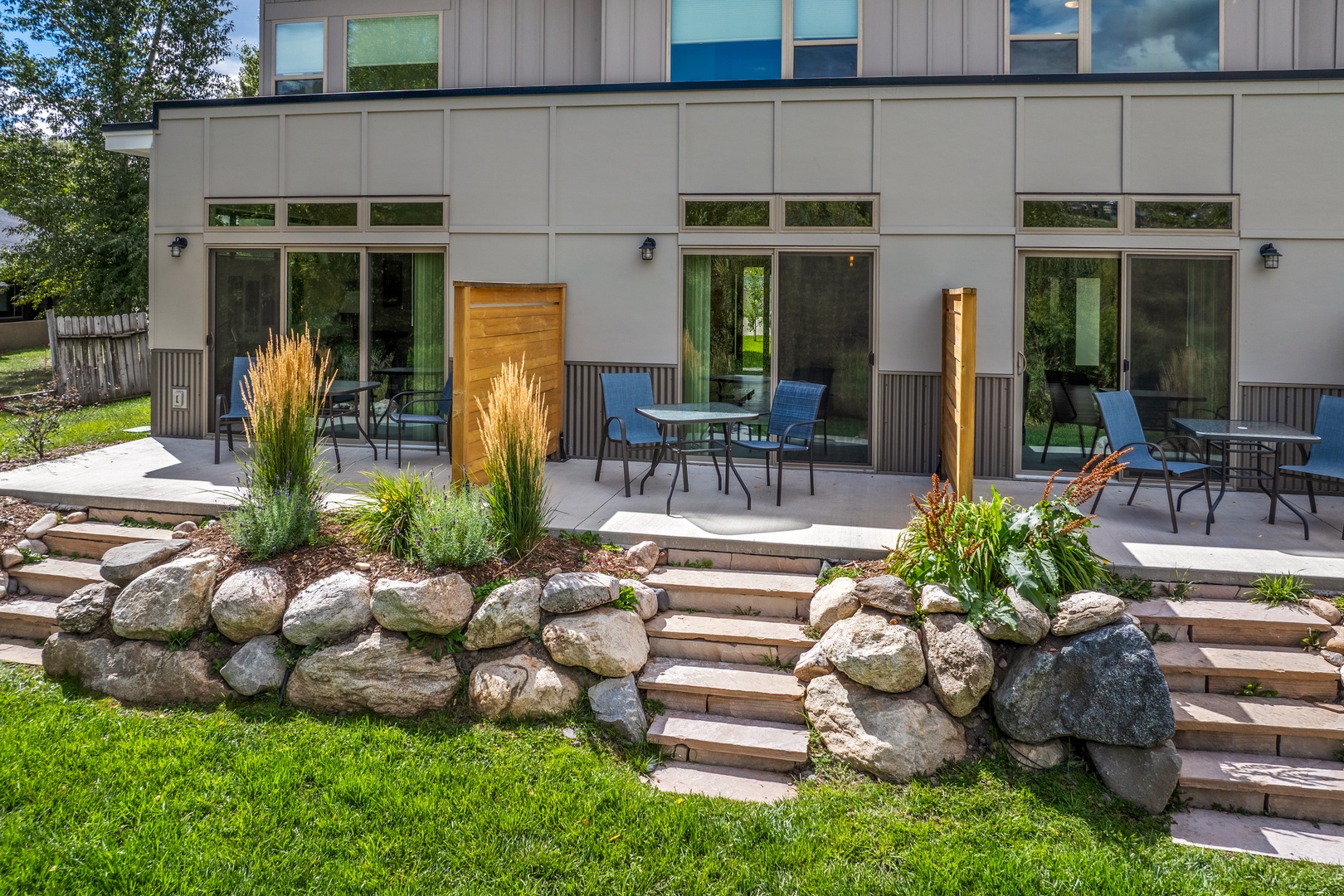 Modern vacation rental exterior features three private patio spaces with seating, separated by privacy screens and bordered by natural stone landscaping, with easy access from ground-level patios and upper decks.