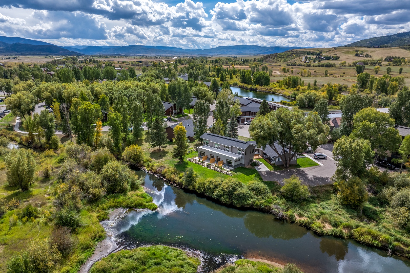 Aerial view of the property nestled along a serene river, surrounded by lush trees and expansive mountain vistas under a dramatic sky.
