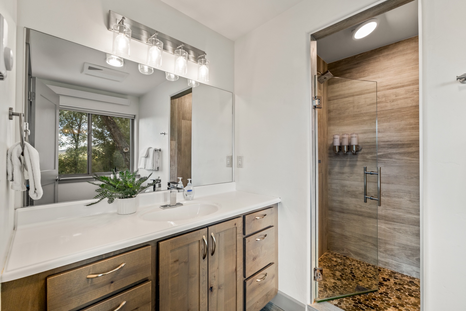 Step into your private spa-like bathroom with a stunning walk-in shower featuring natural wood-look tile, pebble flooring, and a sleek vanity bathed in natural light from your window.