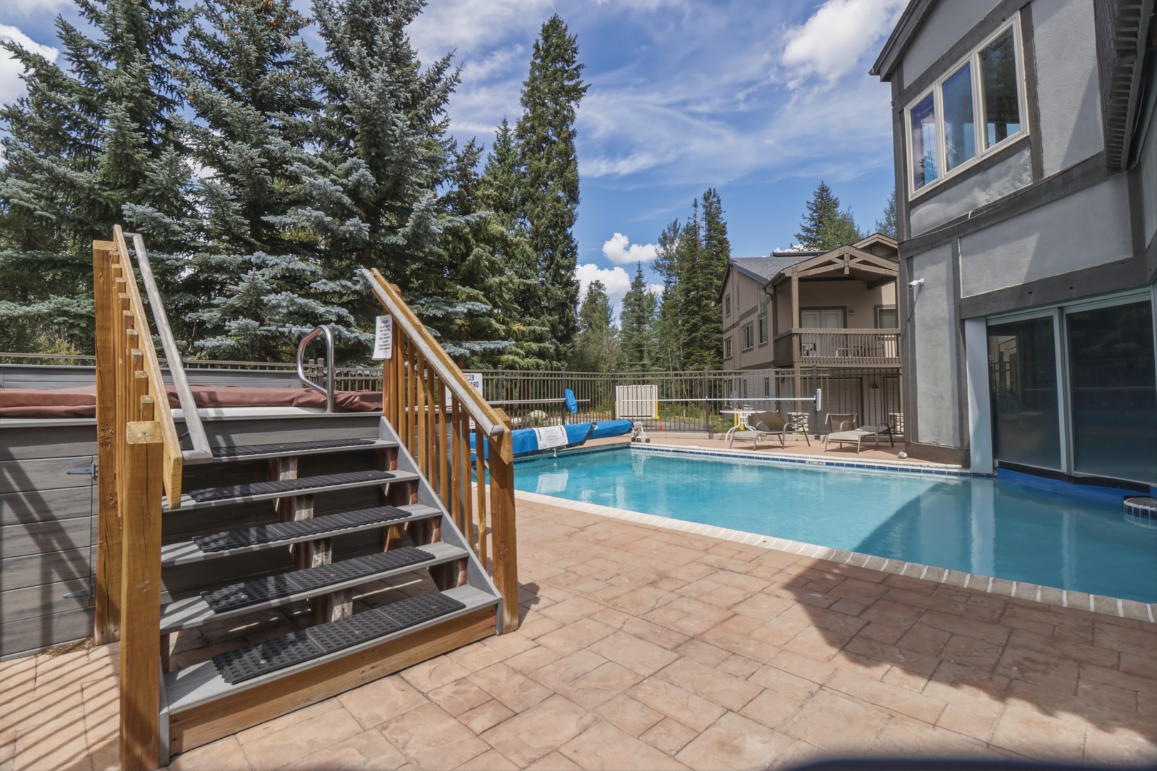 A sparkling pool area surrounded by tall pines, perfect for refreshing mountain getaways. Steps lead to a hot tub, while lounge chairs invite sunny afternoons of relaxation.