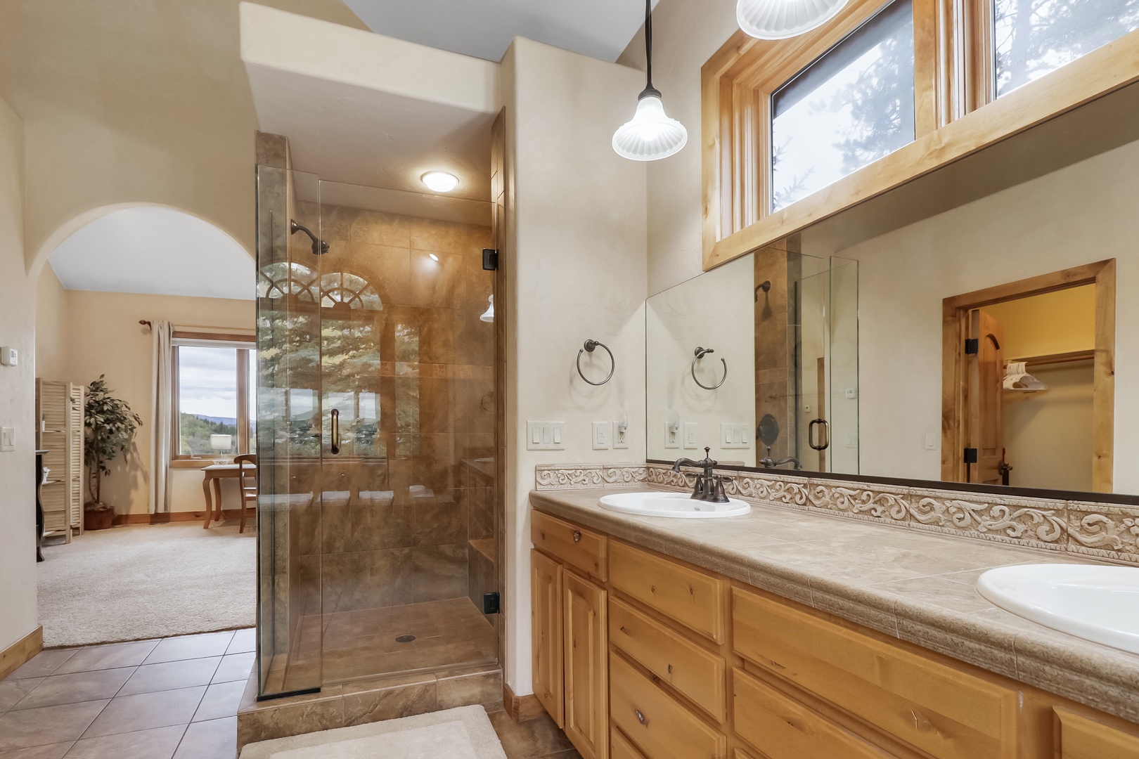 Your spacious bathroom features elegant dual vanities, a large walk-in shower, and abundant natural light streaming through wood-framed windows. The warm tones and thoughtful design create a spa-like retreat.