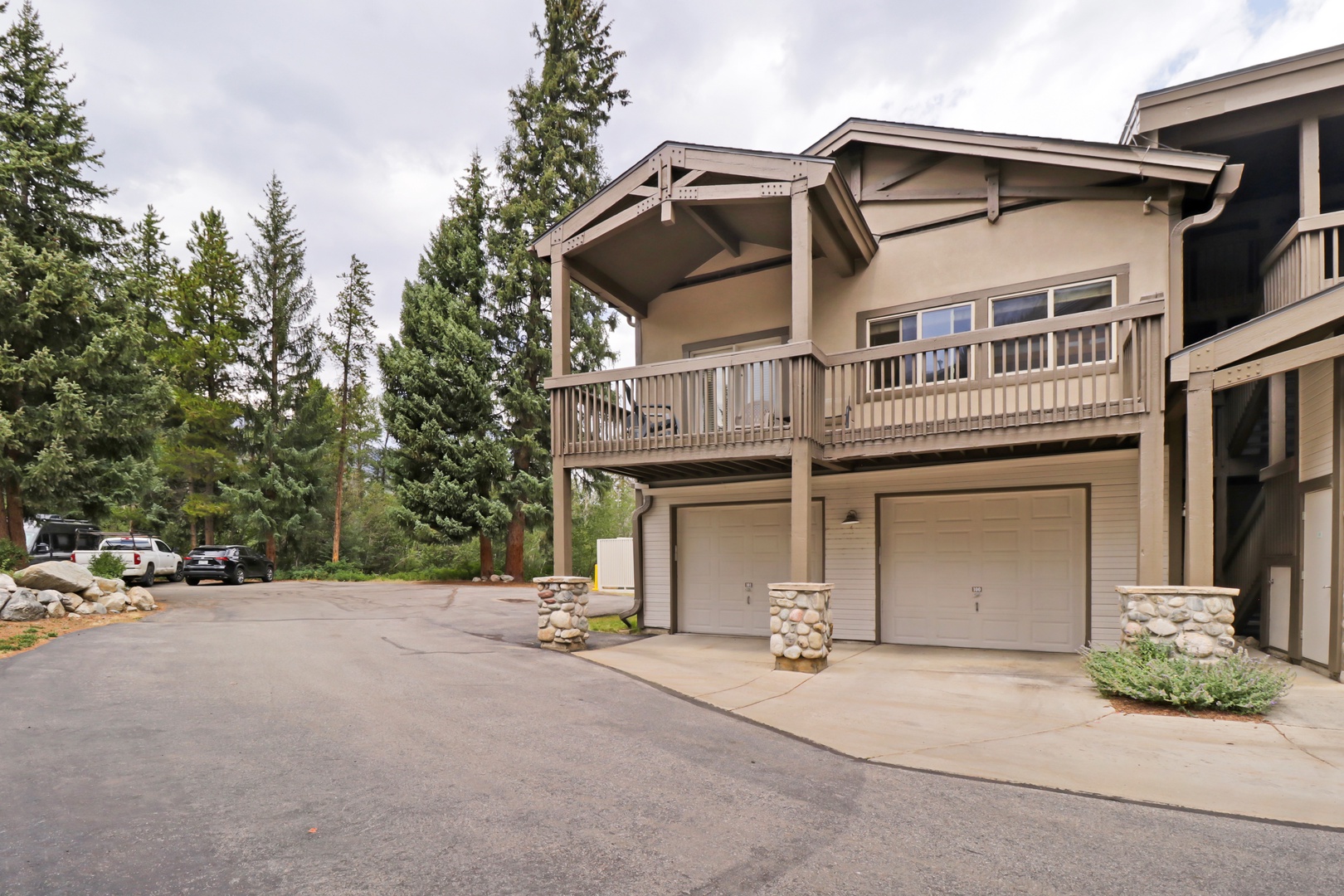 Mountain lodge-style property featuring attached garages and private balcony, nestled among tall evergreens with convenient parking access.
