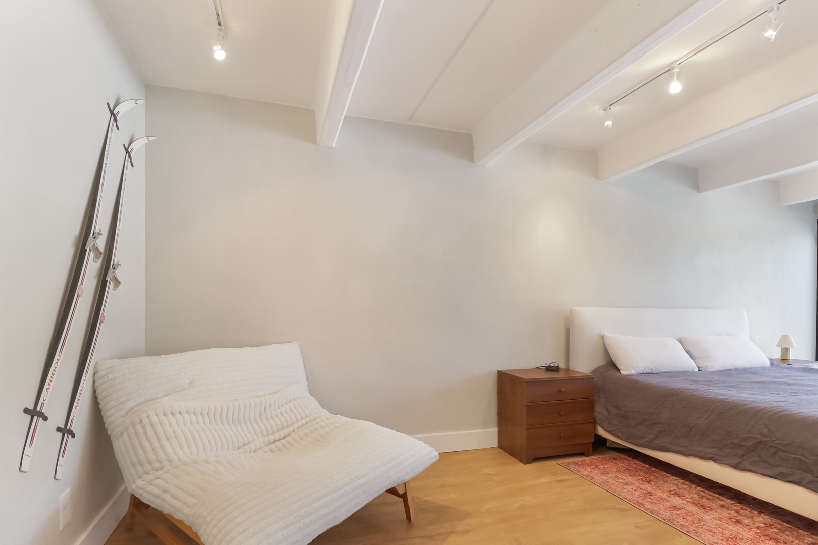 Unwind in your serene bedroom featuring exposed ceiling beams, warm wood flooring, and a cozy reading chair for peaceful moments.