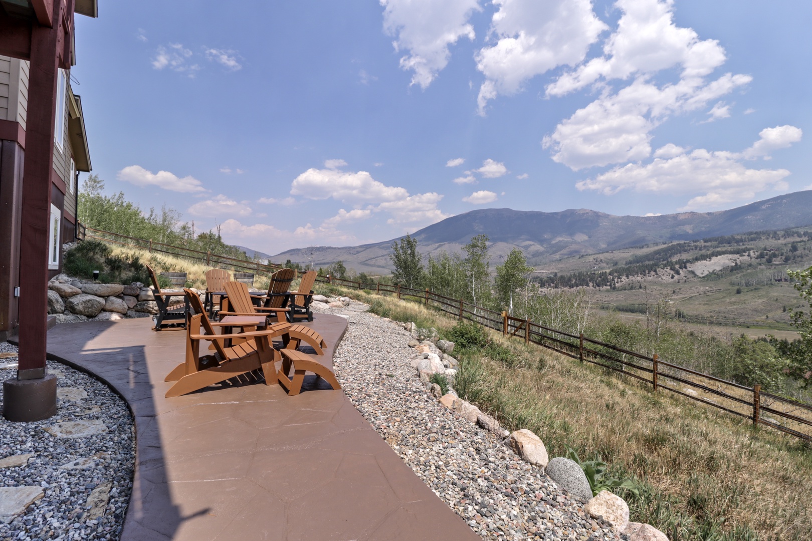 An expansive mountain valley unfolds beyond the property's outdoor seating area, framed by rolling foothills and distant peaks under a brilliant summer sky.