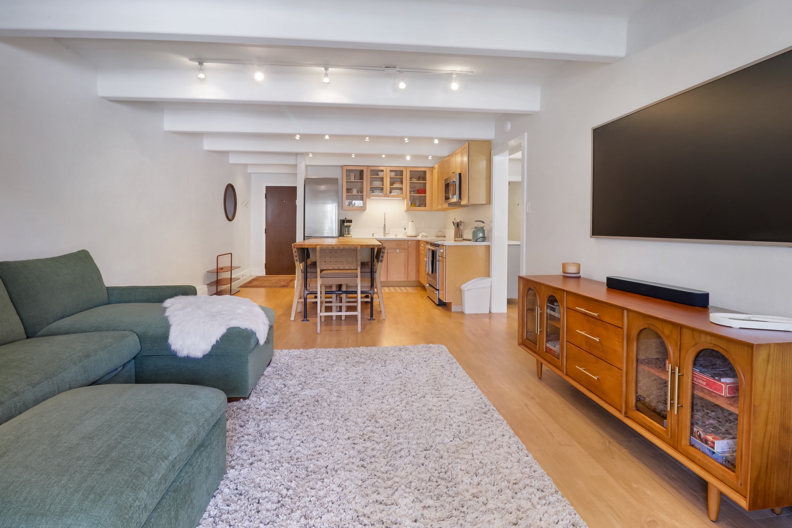 Relax in your stylish open-plan living space featuring a cozy green sofa, warm wood beams, and a fully equipped kitchen.