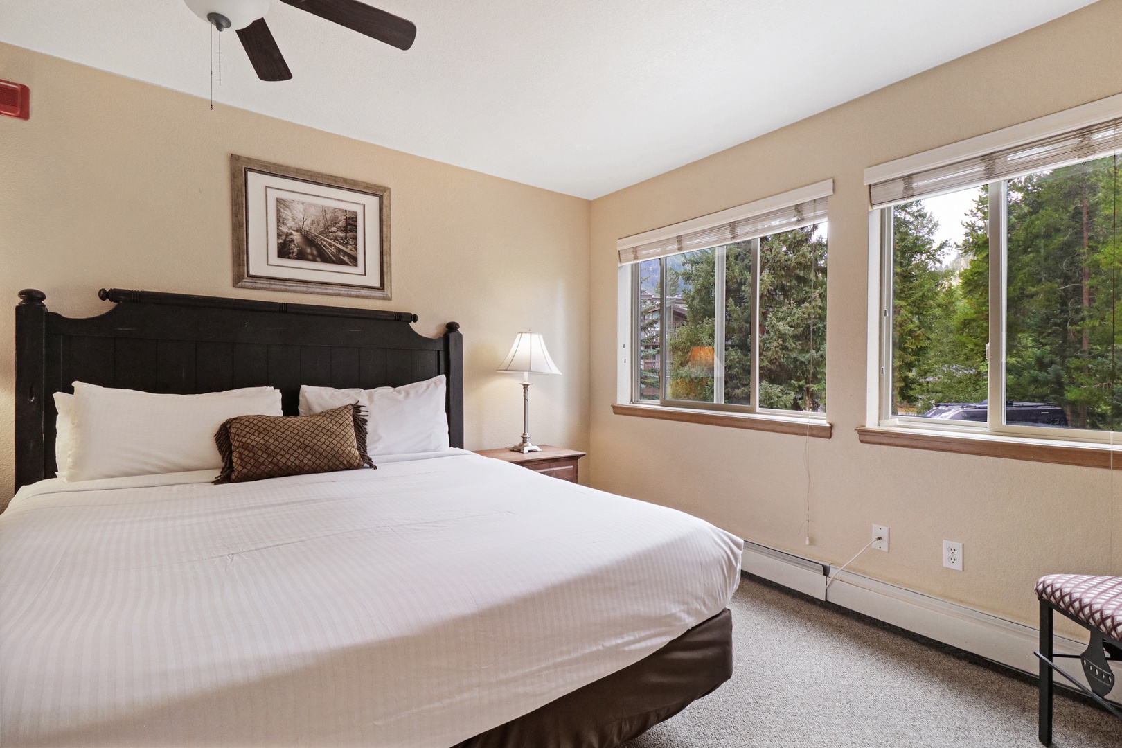 Rest peacefully in your private bedroom with plush white bedding, warm wood accents, and windows framing lush forest views. The gentle ceiling fan ensures your comfort throughout the night.