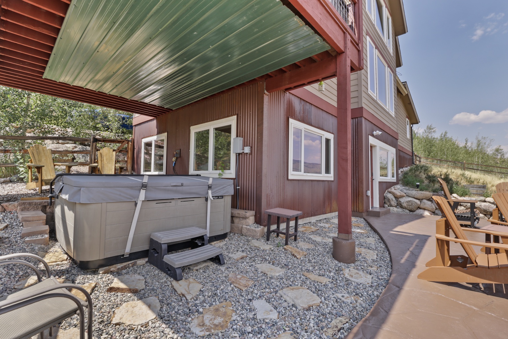 A covered patio area with a private hot tub, stone pathways, wooden seating, and open views of the surrounding natural landscape perfect for outdoor relaxation.