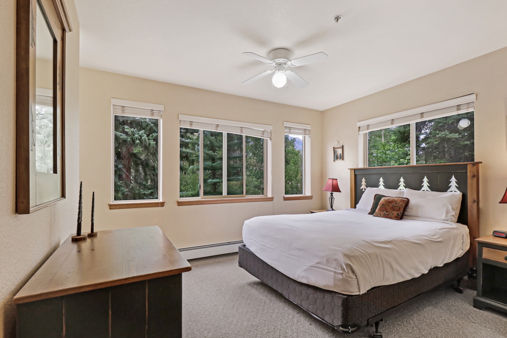 Drift off in your serene bedroom with multiple windows framing the surrounding evergreens, creating a peaceful mountain retreat ambiance as natural light fills the space.
