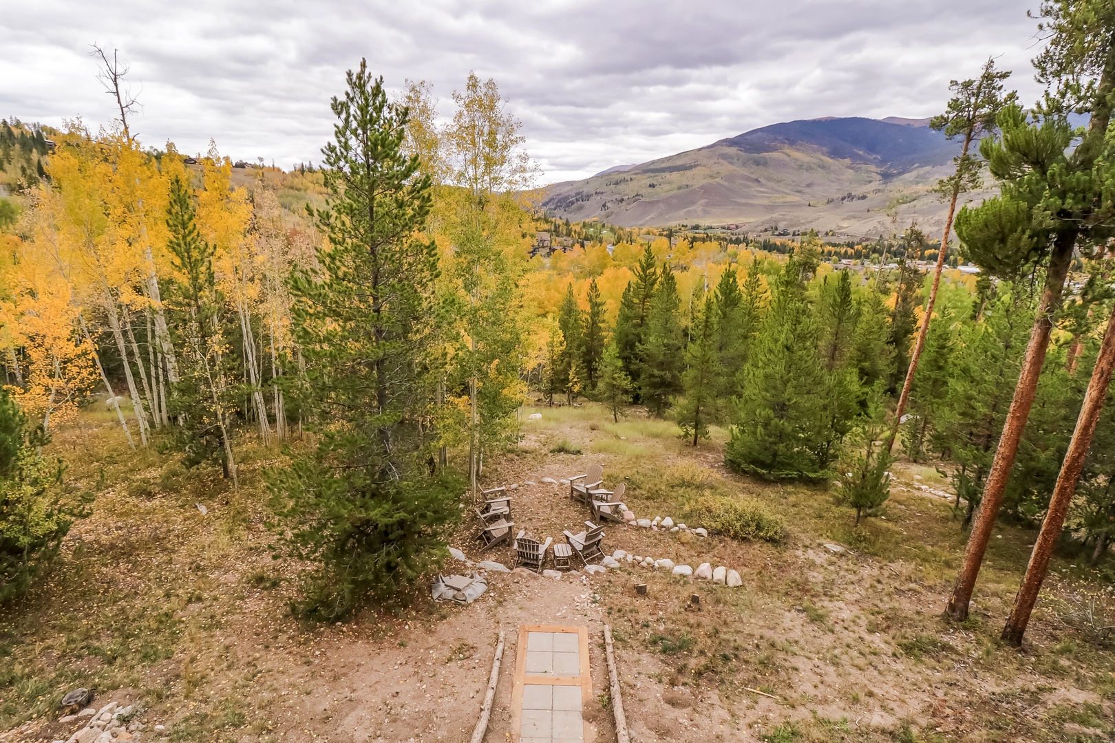 An elevated property view showcasing a private outdoor fire pit surrounded by aspens in golden autumn foliage, evergreens, and dramatic mountain scenery in the distance.