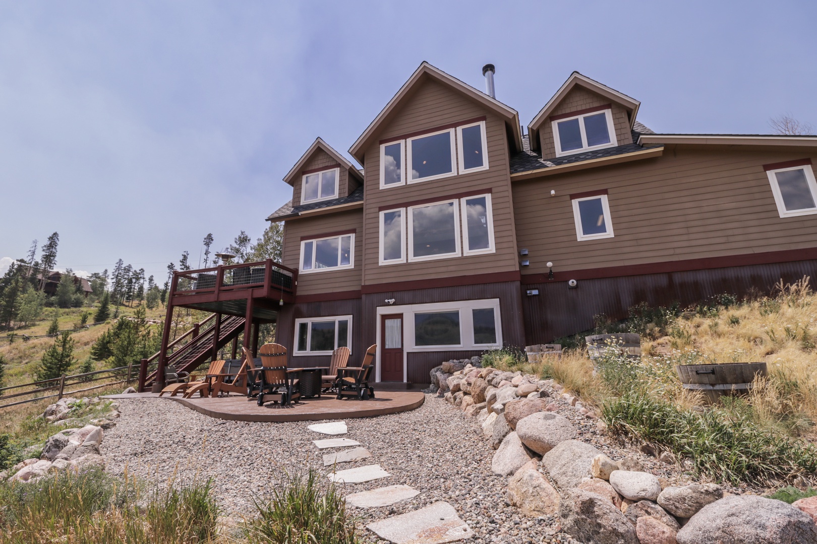 A spacious mountain retreat set on a gentle hillside, featuring multiple levels of outdoor gathering spaces. The patio invites you to relax around the fire ring while taking in the surrounding natural landscape and seasonal greenery.