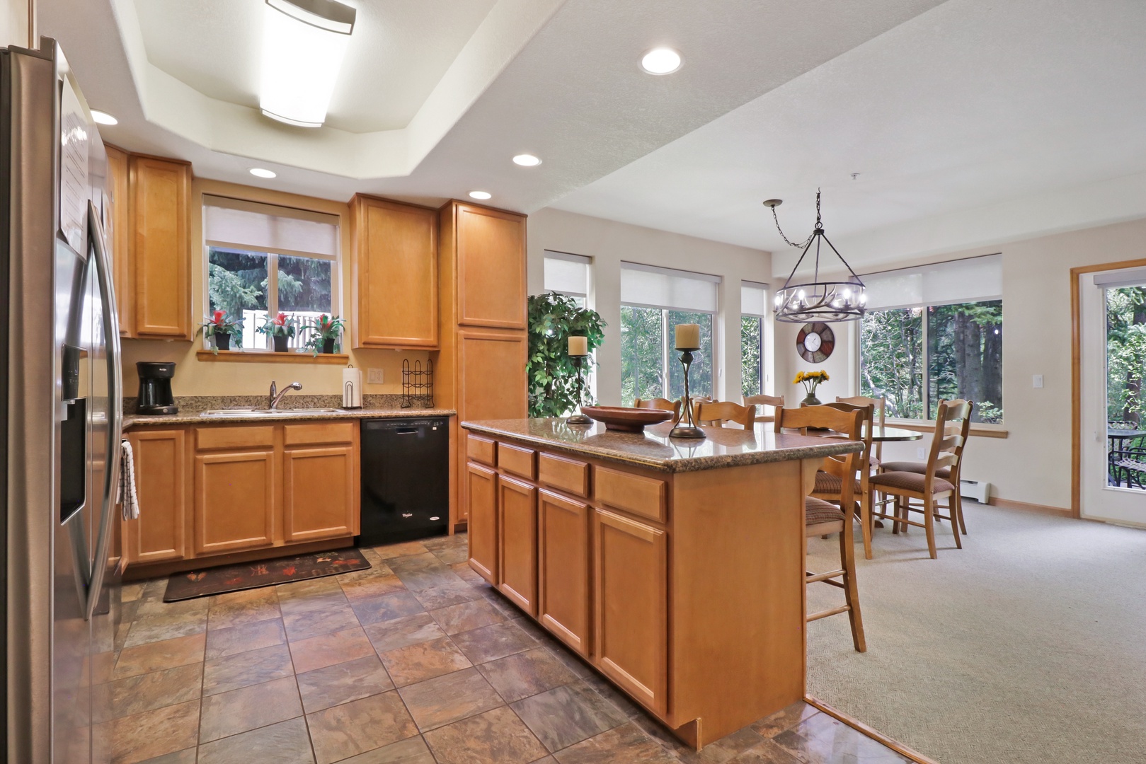 Gather around the spacious kitchen island with granite counters and warm wood cabinetry. Large windows frame peaceful forest views, flooding your culinary space with natural light.